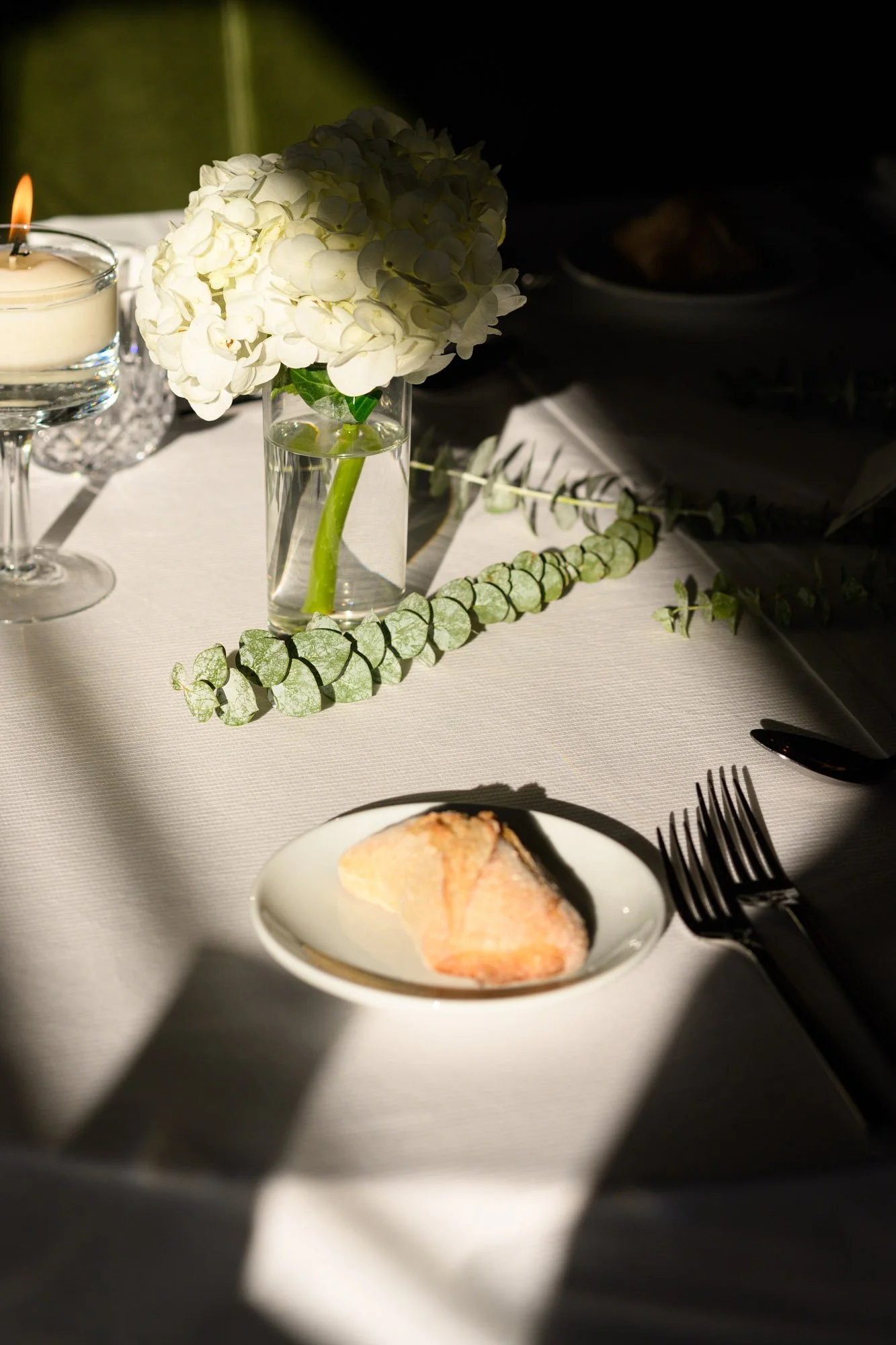 Table élégante avec une nappe blanche, une bougie, un vase avec des hortensias blancs, une guirlande de feuilles, une assiette avec une pâtisserie, une fourchette et un couteau.