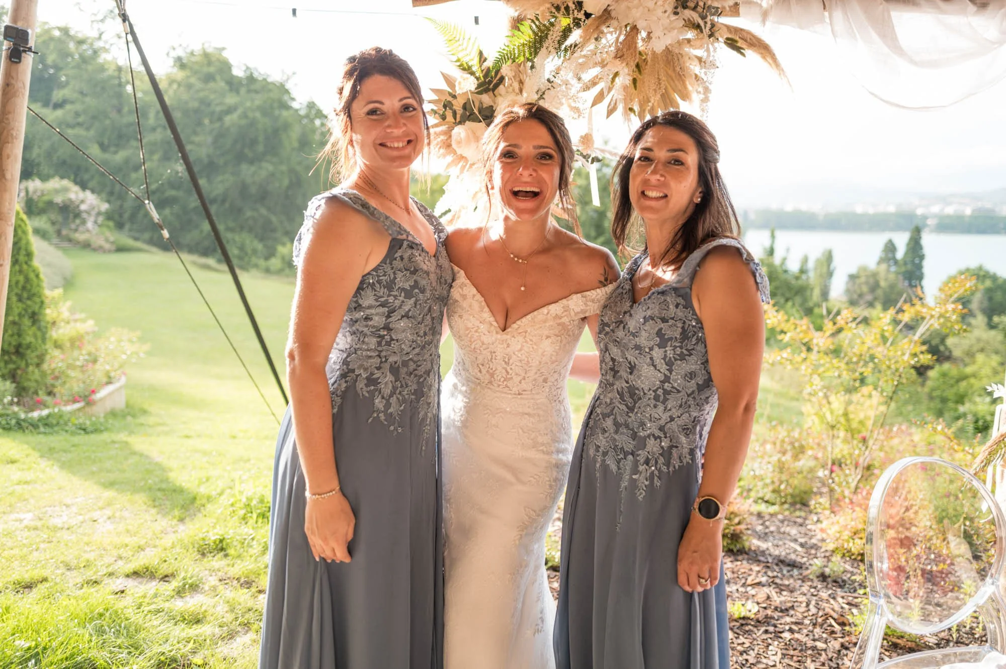 Trois femmes posent en extérieur, une dans une robe de mariée et deux en robes longues gris avec des détails en dentelle, lors d'une célébration de mariage.