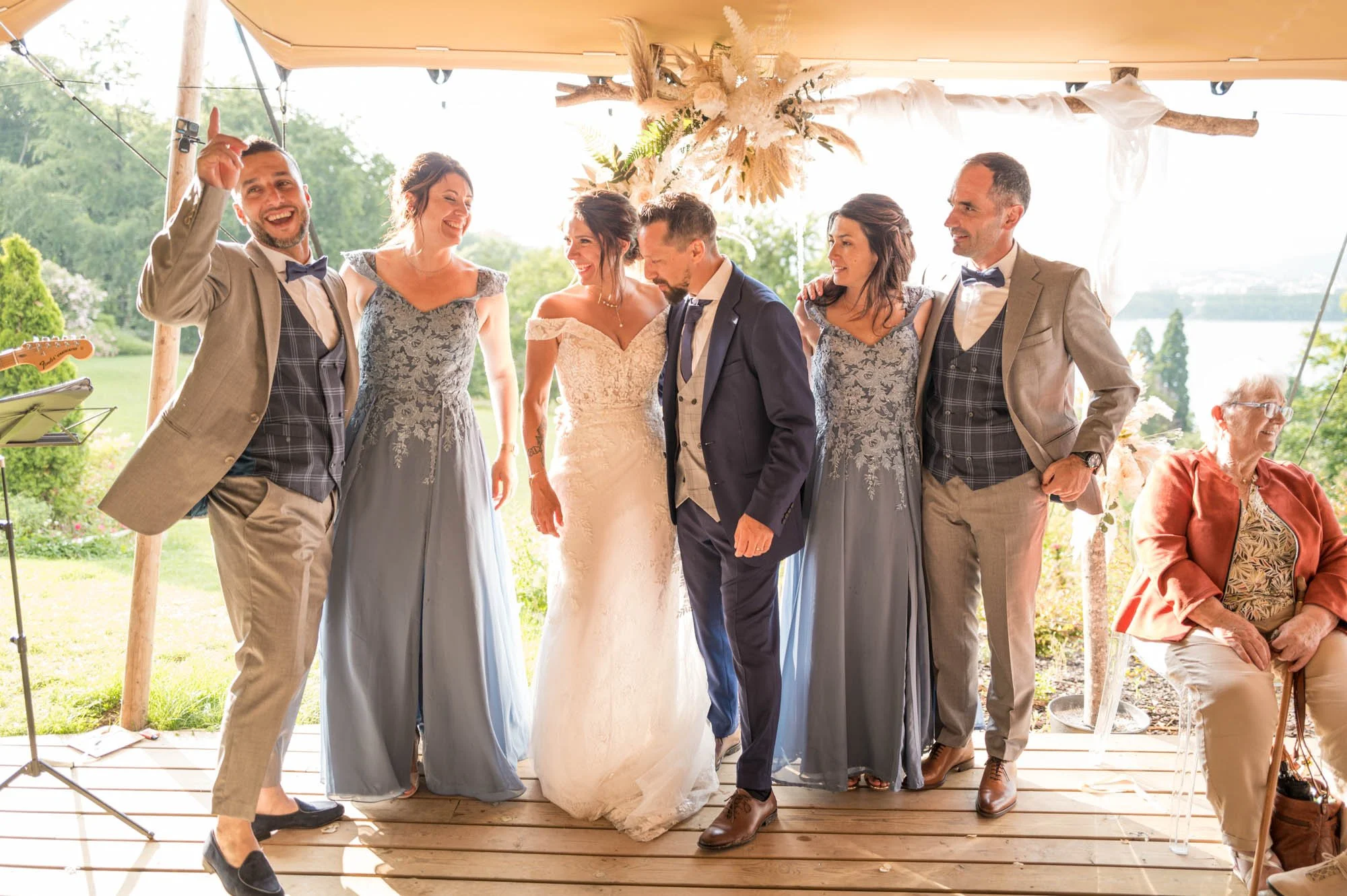 Groupe de personnes lors d'un mariage, avec la mariée en robe blanche, le marié en costume, et quatre autres personnes en robes et costumes assortis, sous une tente avec un décor naturel en arrière-plan.
