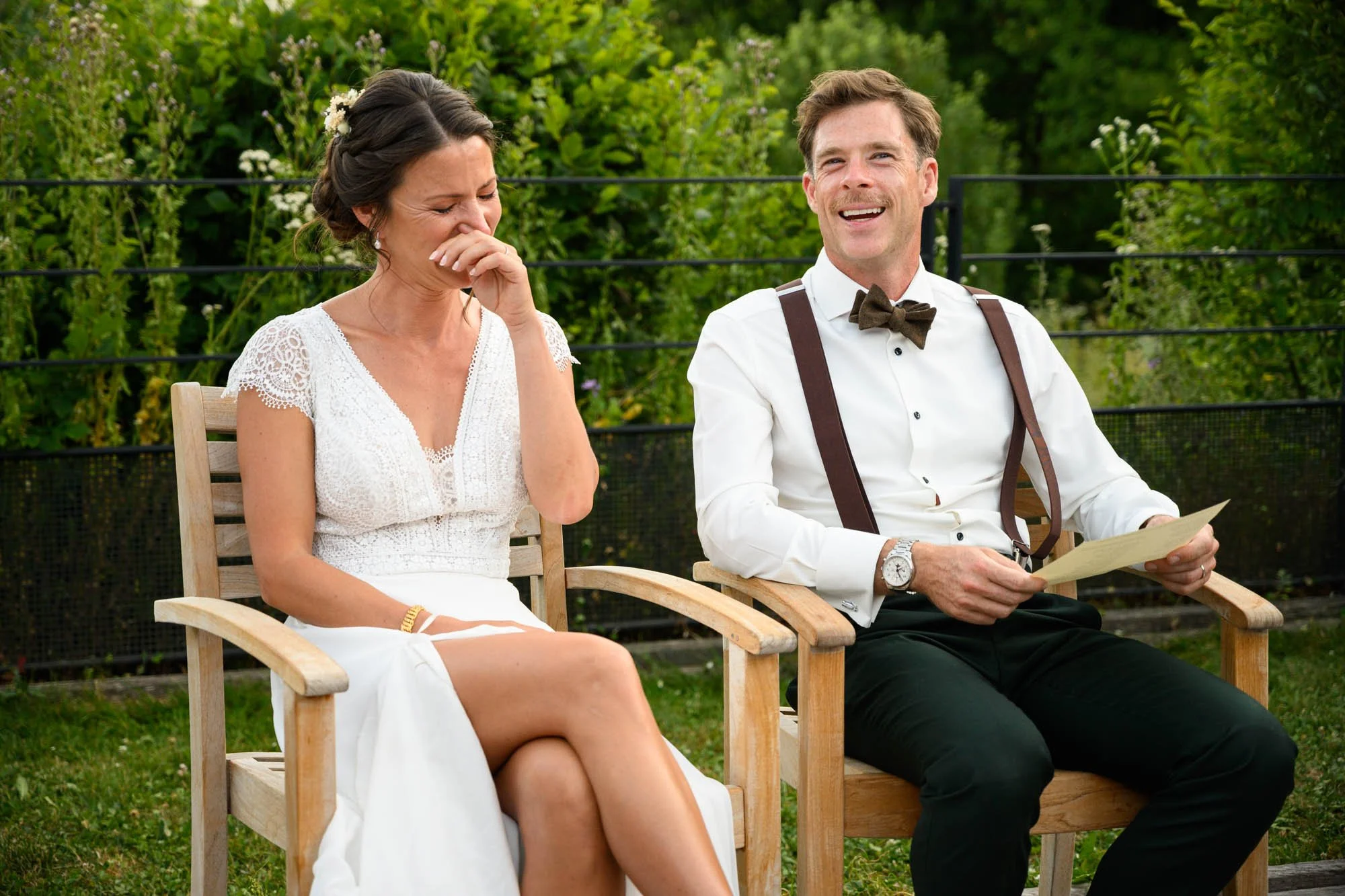 Une femme et un homme assis dehors, tous deux souriant et riant, lors d'une cérémonie en plein air. La femme porte une robe blanche avec des détails en dentelle, et l'homme porte une chemise blanche avec un nœud papillon et des bretelles.