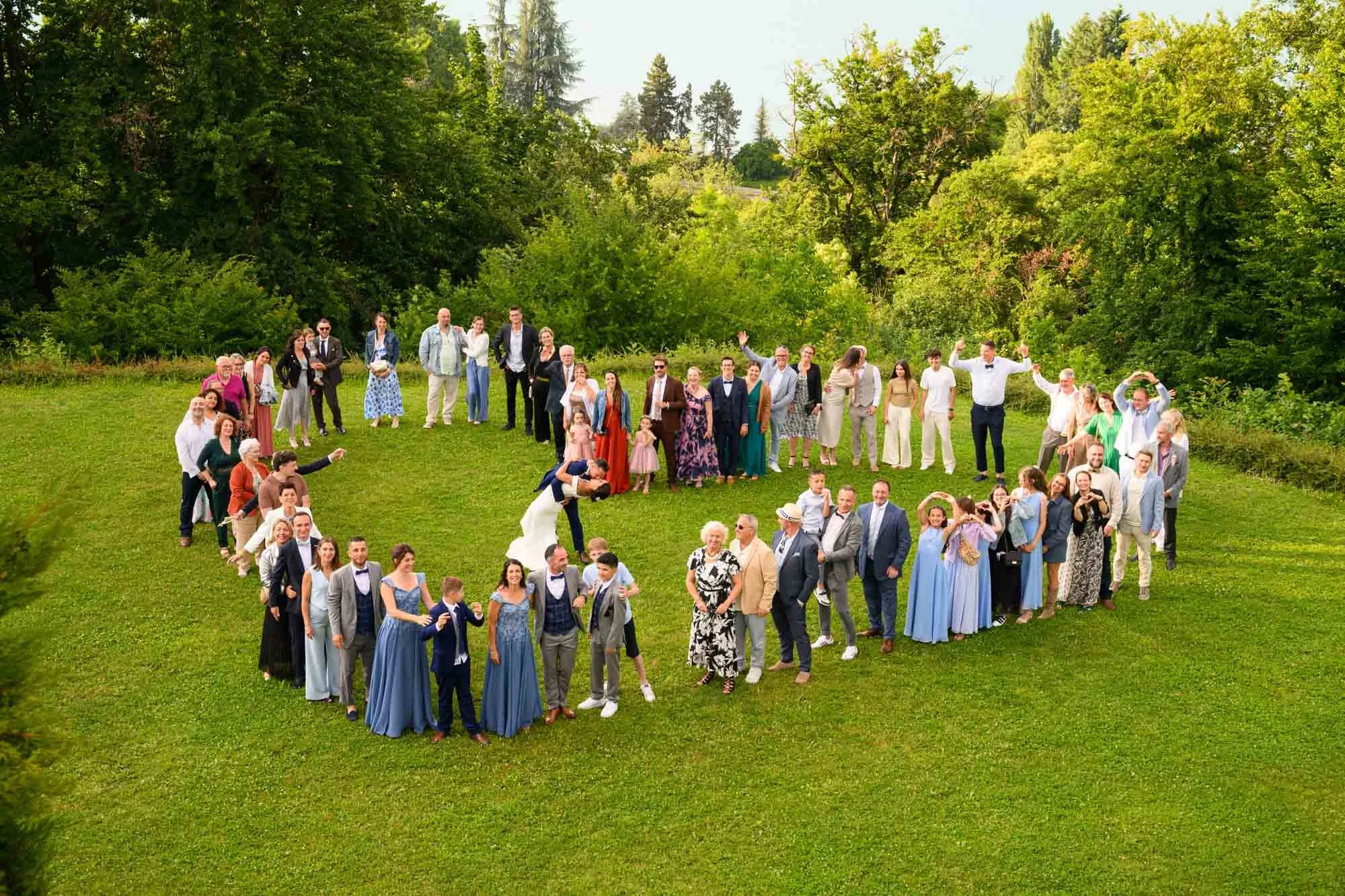 groupe de personnes lors d'un mariage en plein air, formant un cœur avec l'herbe verte et les arbres en arrière-plan.