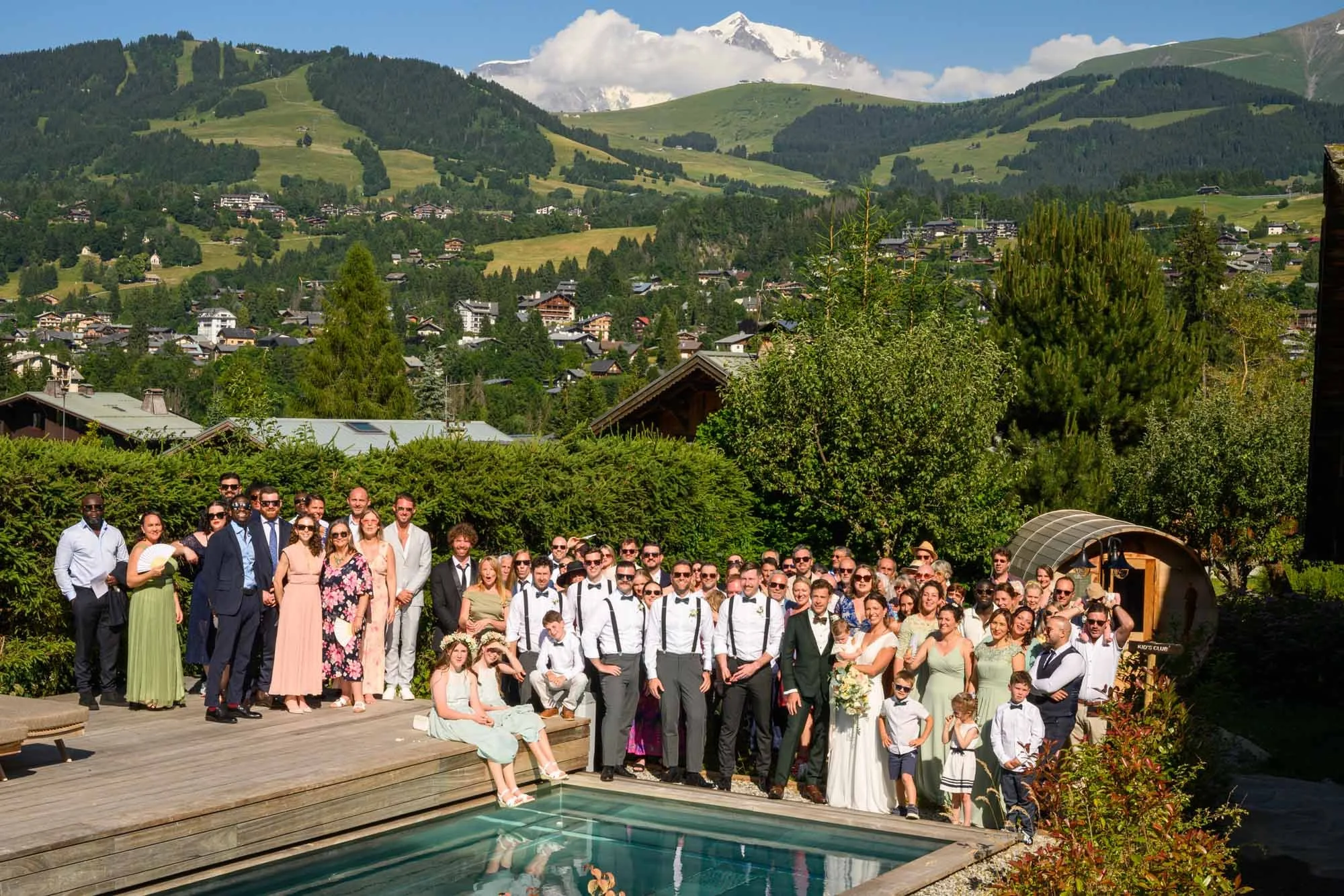 groupe de personnes lors d'un mariage en plein air à côté d'une piscine, avec un fond de montagnes verdoyantes et de maisons dans une vallée
