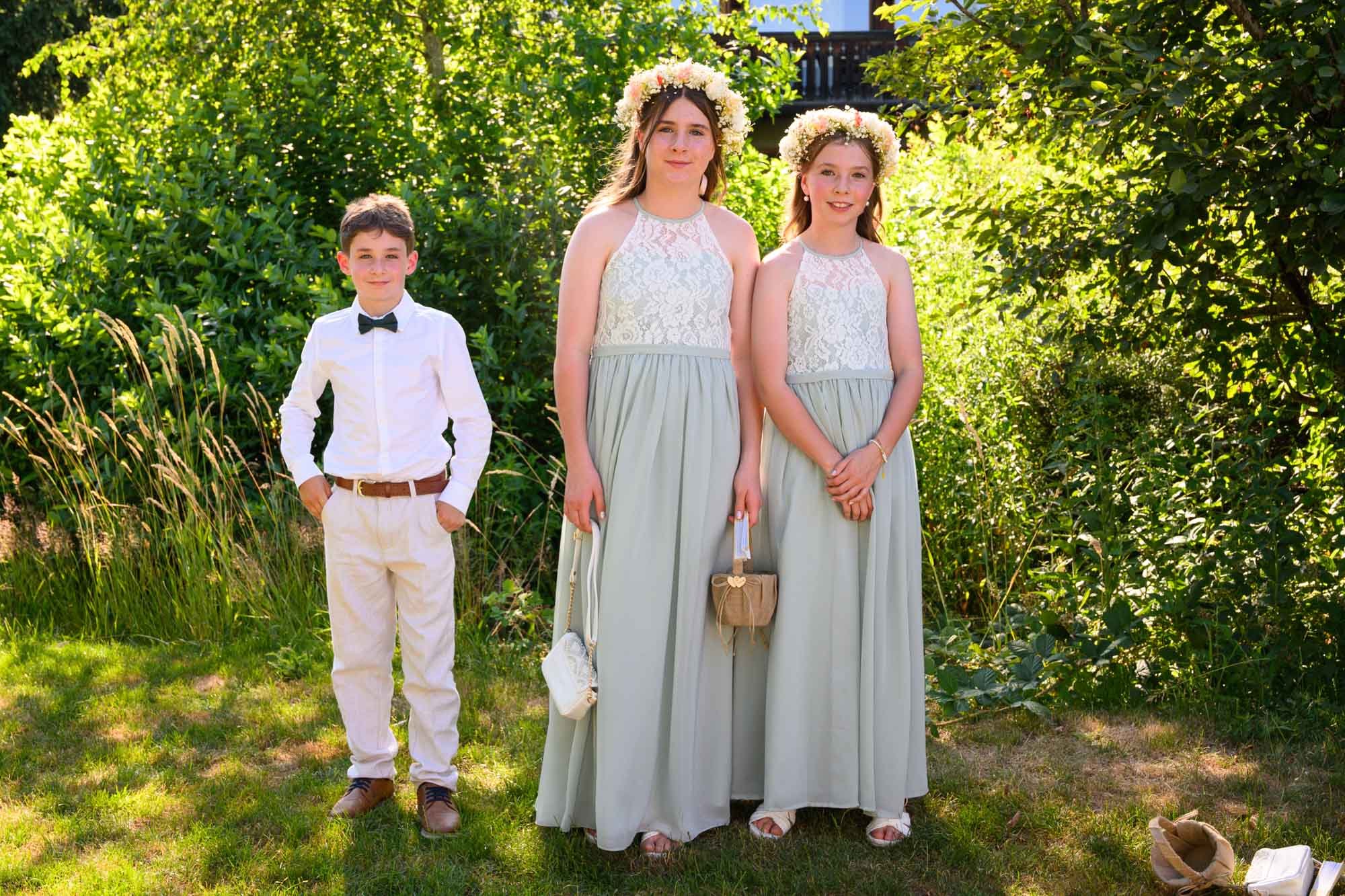 Trois enfants en vêtements formels posent devant un fond de verdure. Les deux filles portent des robes longues gris clair avec des couronnes de fleurs, et le garçon porte une chemise blanche avec un nœud papillon, un pantalon beige et des chaussures 