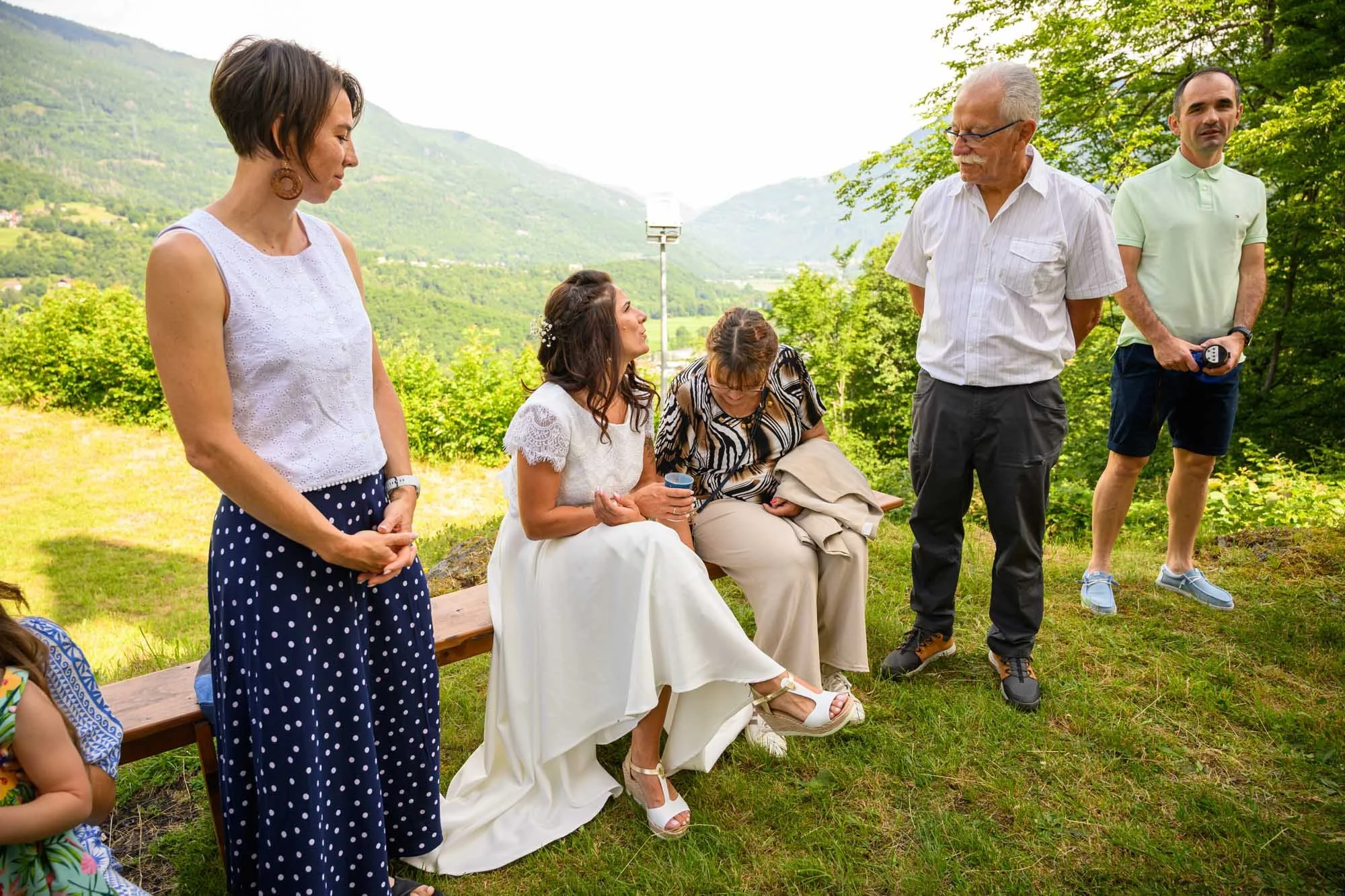 Groupe de personnes lors d'une cérémonie en plein air dans un environnement verdoyant, avec montagnes en arrière-plan. Une femme en robe blanche, appuyée sur une chaise, semble être la mariée. Autour d'elle, plusieurs personnes discutent ou regardent