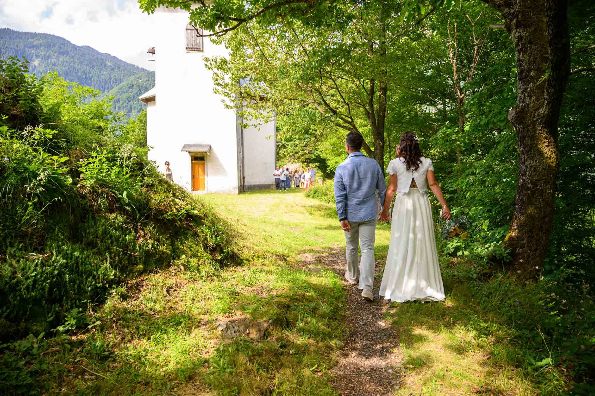 Un couple marche main dans la main sur un sentier forestier vers une église blanche, avec des invités en arrière-plan, dans un cadre verdoyant.