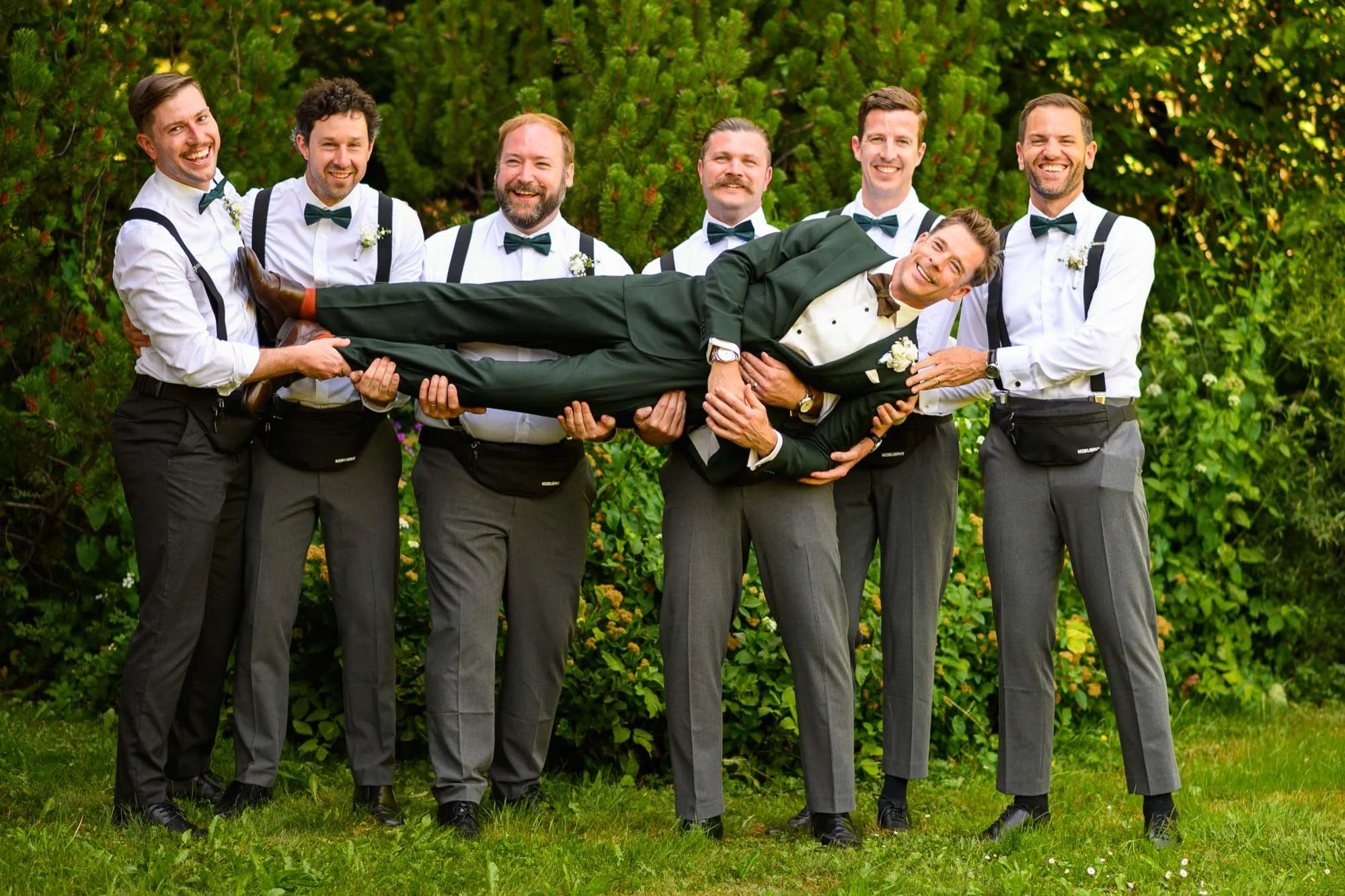 Un groupe d'hommes en tenue de mariage, souriants, où cinq d'entre eux tiennent un homme vêtu en costume de mariage allongé horizontalement, en pleine nature.