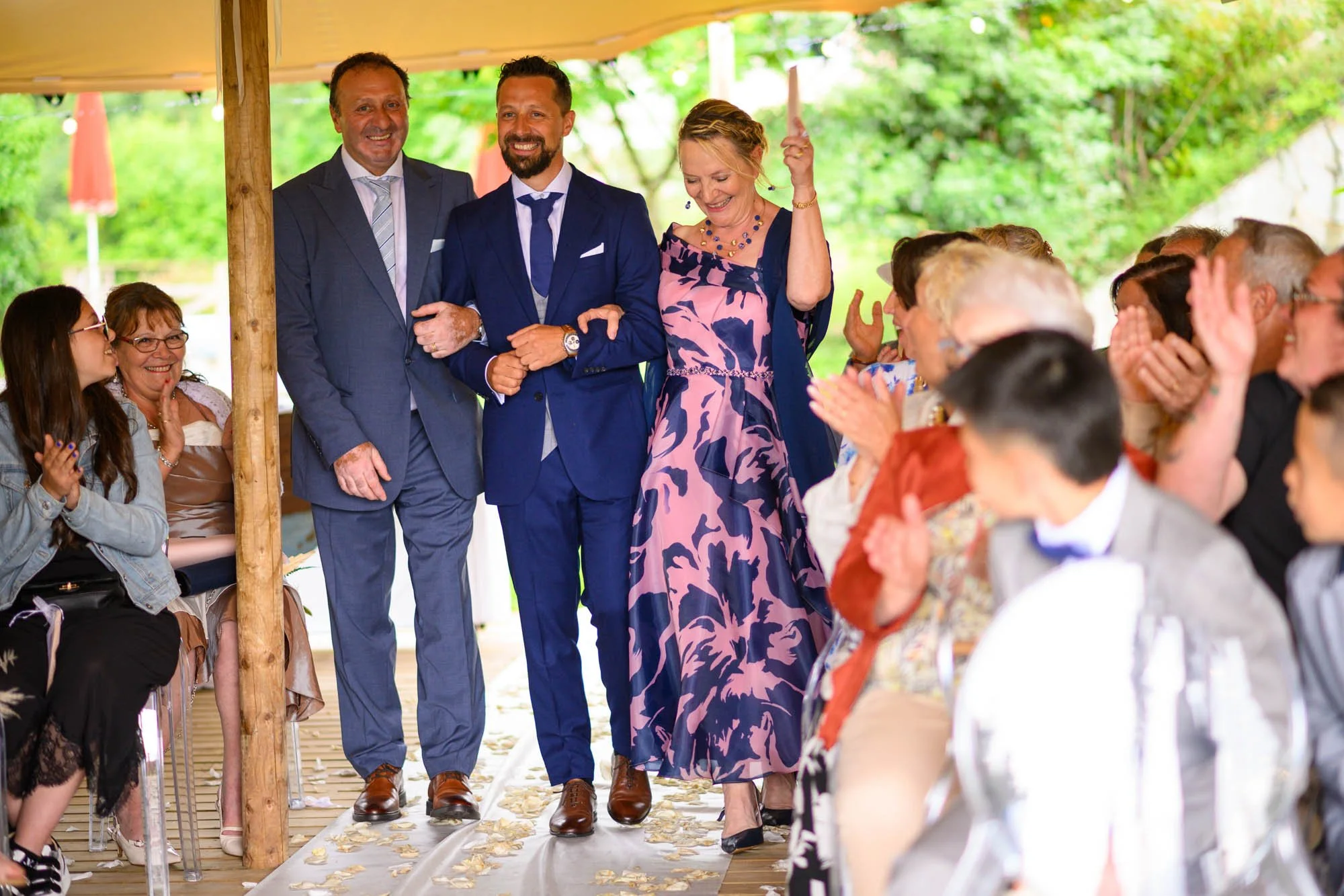 Groupe de personnes à un mariage, certains personnes assises applaudissent, des invités en arrière-plan. Troisième personne en partant de la gauche, un homme en costume bleu marche dans l'allée en tenant la main d'une femme en robe rose et bleue. Aut