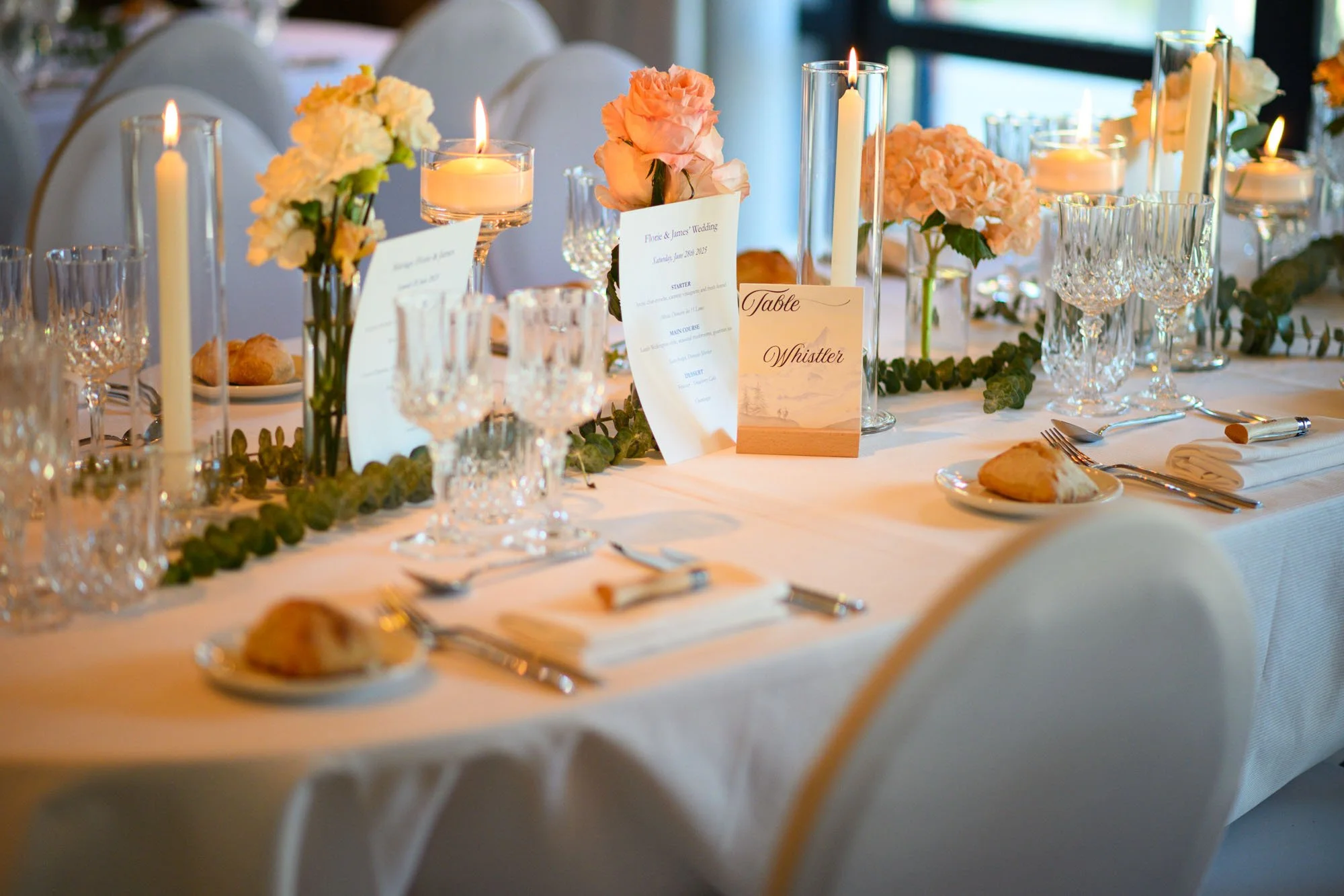 Table de mariage élégante avec nappes blanches, centres de fleurs roses et blanches, bougies et vaisselle en cristal. Signes indiquant 'Table Whistler' et une carte de menu.