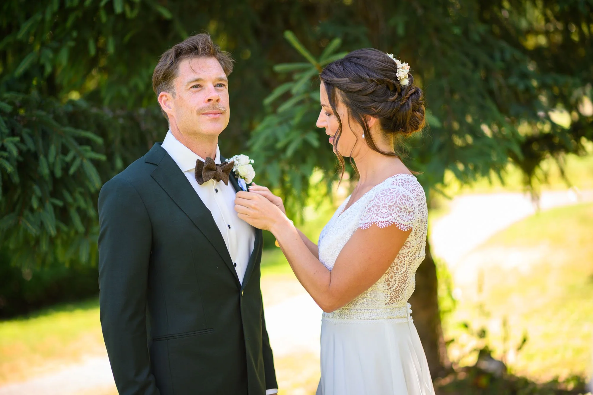 Un couple en costume de mariage, un homme en smoking et une femme en robe blanche, dans un cadre extérieur avec des arbres.