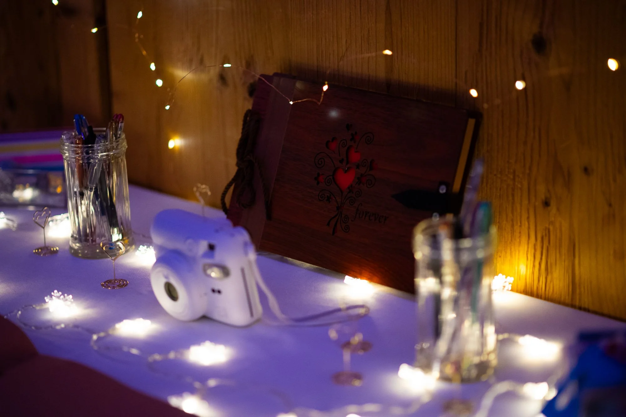 Livre en bois avec des cœurs rouges et la phrase « forever » posé sur une table décorée de guirlandes lumineuses. Quelques vases en verre contenant des stylos et un appareil photo blanc à côté de l'ouvrage.