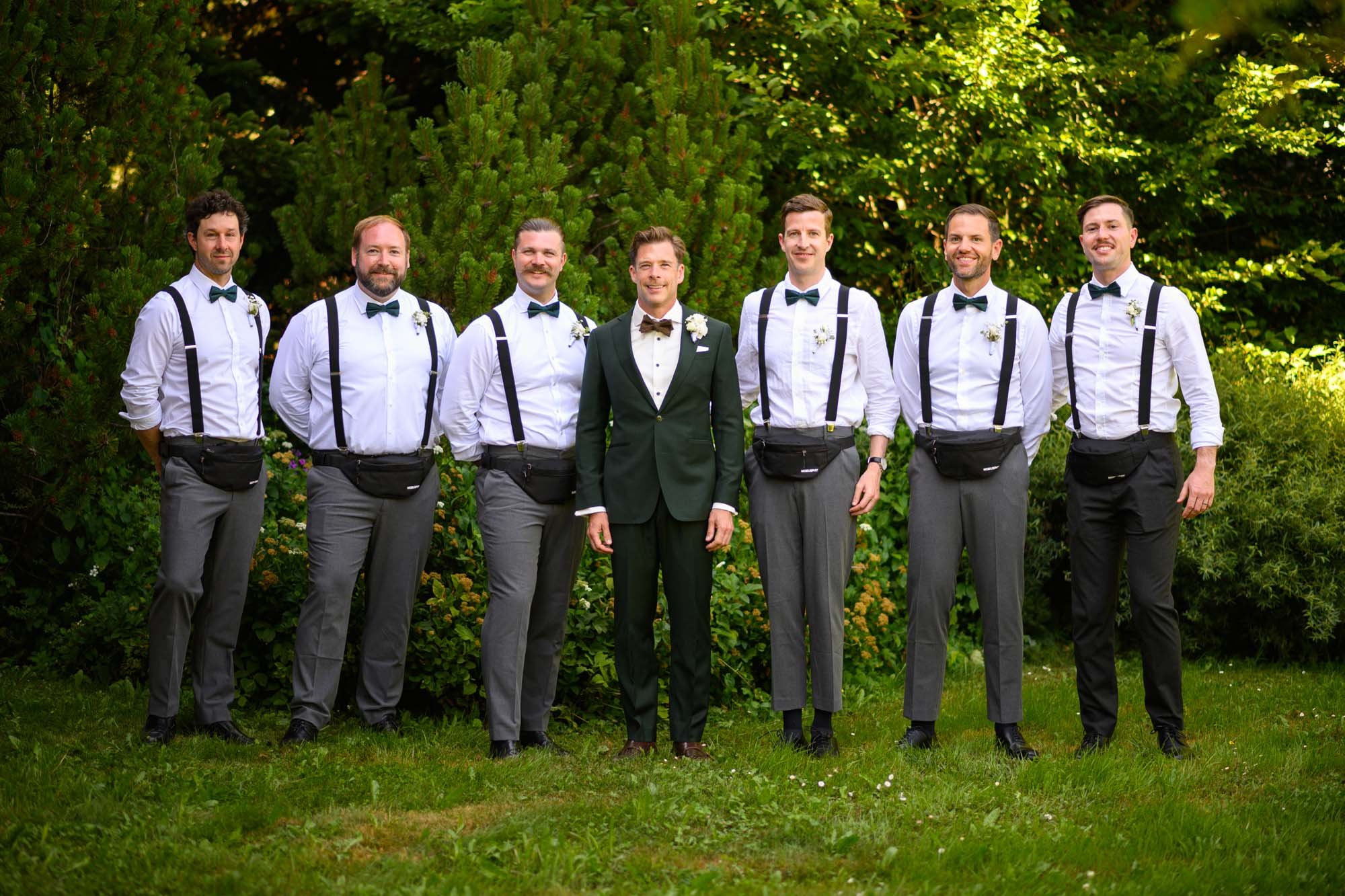 Groupe d'hommes en tenue formelle, dont le marié au centre en costume vert, lors d'un mariage en extérieur avec végétation luxuriante en arrière-plan.