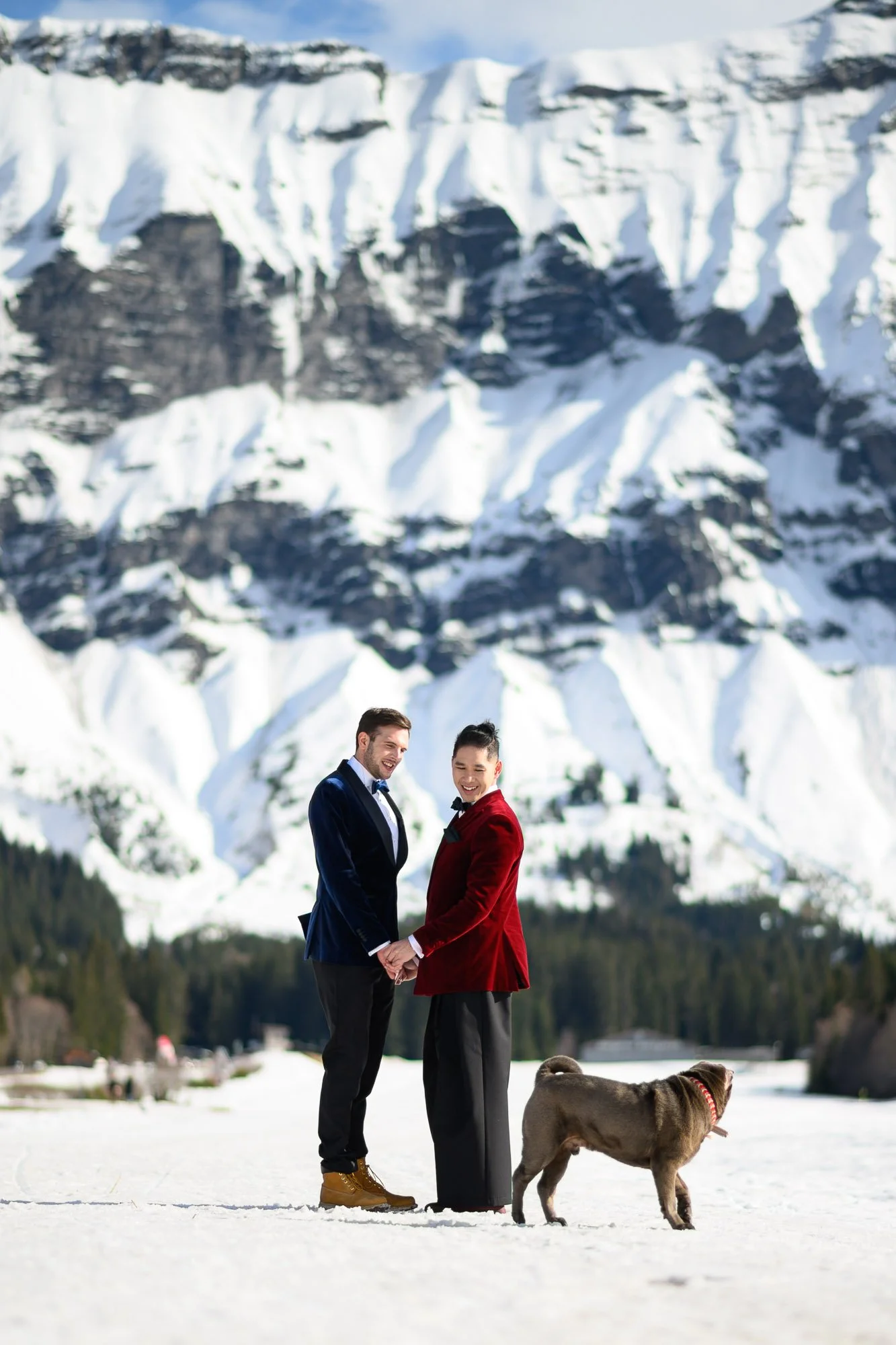 Deux personnes habillées en manteaux de cérémonie, tenant la main, avec un chien dans un paysage enneigé et montagneux en arrière-plan.