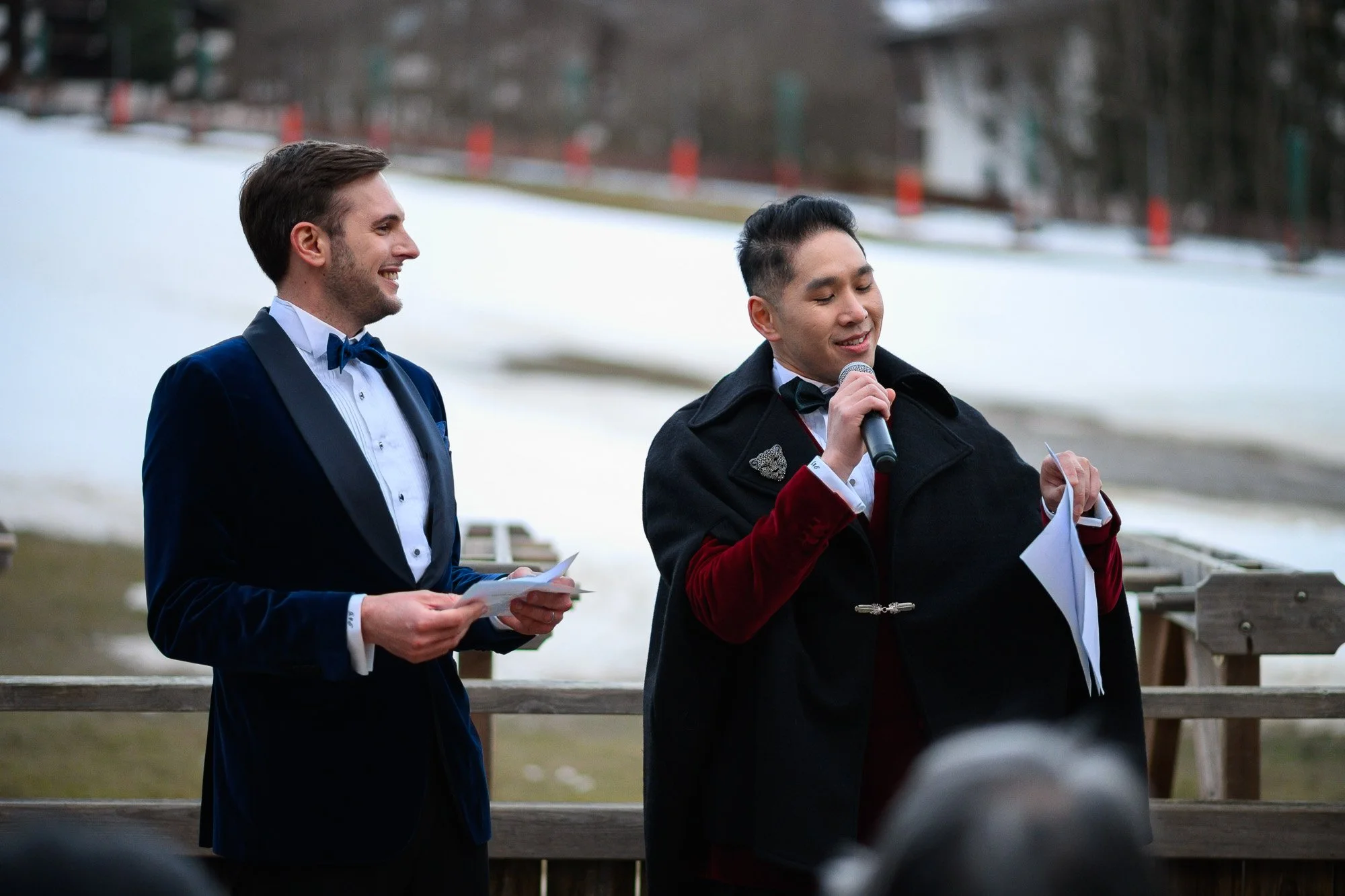 Deux hommes habillés formellement lors d'une cérémonie à l'extérieur, l'un d'eux tenant un micro et des papiers, en arrière-plan une pente enneigée.