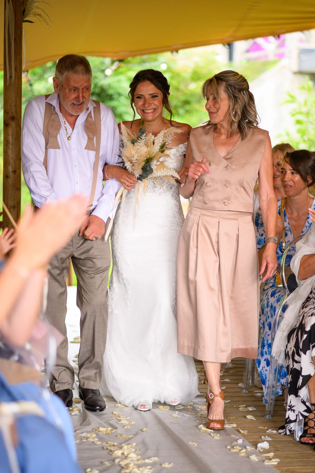 Une femme en robe de mariage souriant, accompagnée de deux personnes, lors d'un mariage en plein air, avec des invités assis et des pétales de fleurs au sol.
