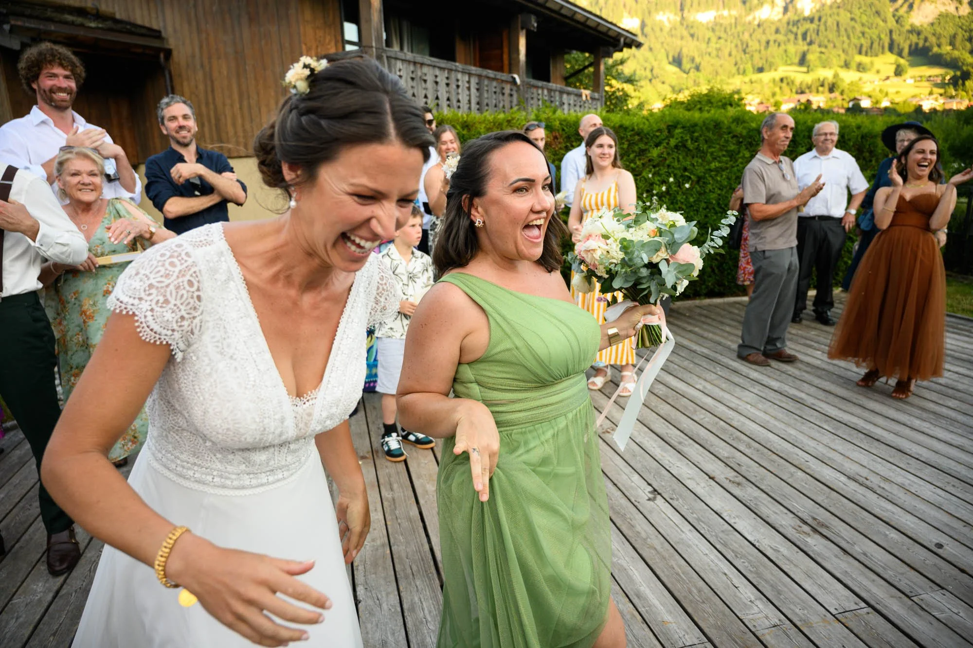 Deux femmes souriantes et heureuses lors d'un mariage en plein air. L'une porte une robe blanche avec des détails en crochet, l'autre porte une robe verte et tient un bouquet de fleurs. Elles semblent danser ou célébrer joyeusement, entourées d'invit