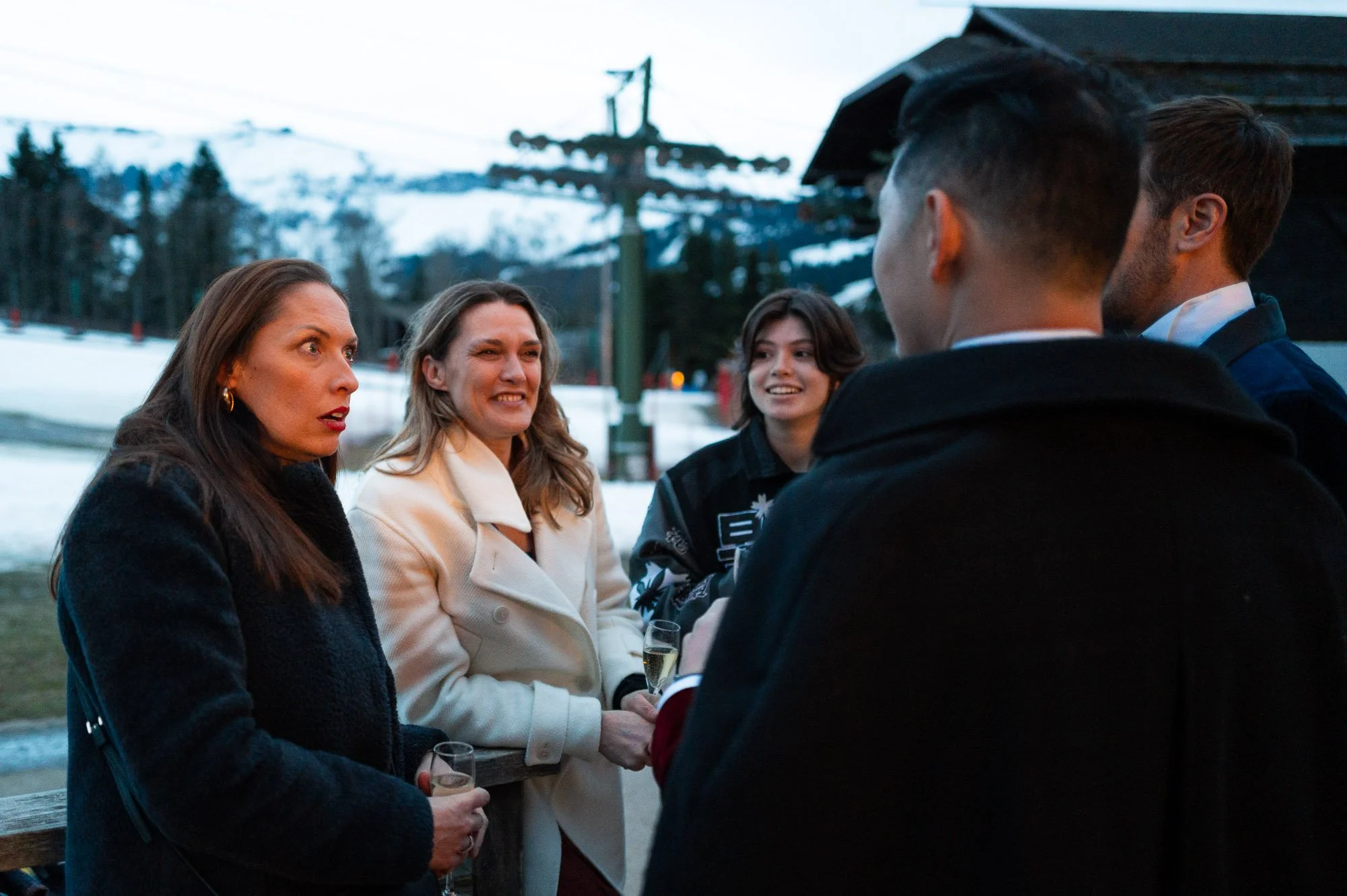 Groupe de cinq personnes, deux femmes et trois hommes, discutant en plein air avec un décor de montagne enneigée en arrière-plan, lors d'une soirée.