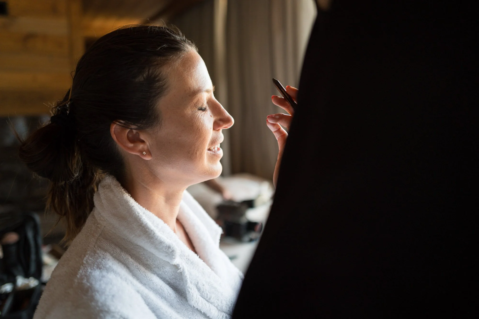 Une femme souriante en peignoir blanc, les yeux fermés, recevant probablement un soin ou maquillage, avec un fond intérieur chaleureux.
