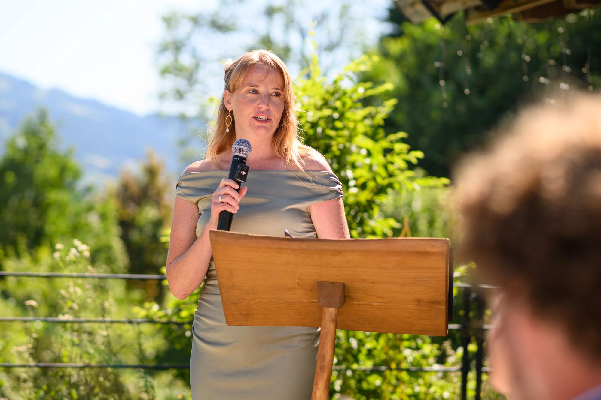Une femme habillée d'une robe moulante couleur or parle en tenant un micro lors d'une présentation en extérieur, avec un pupitre en bois devant elle, entourée de végétation et de montagnes en arrière-plan.