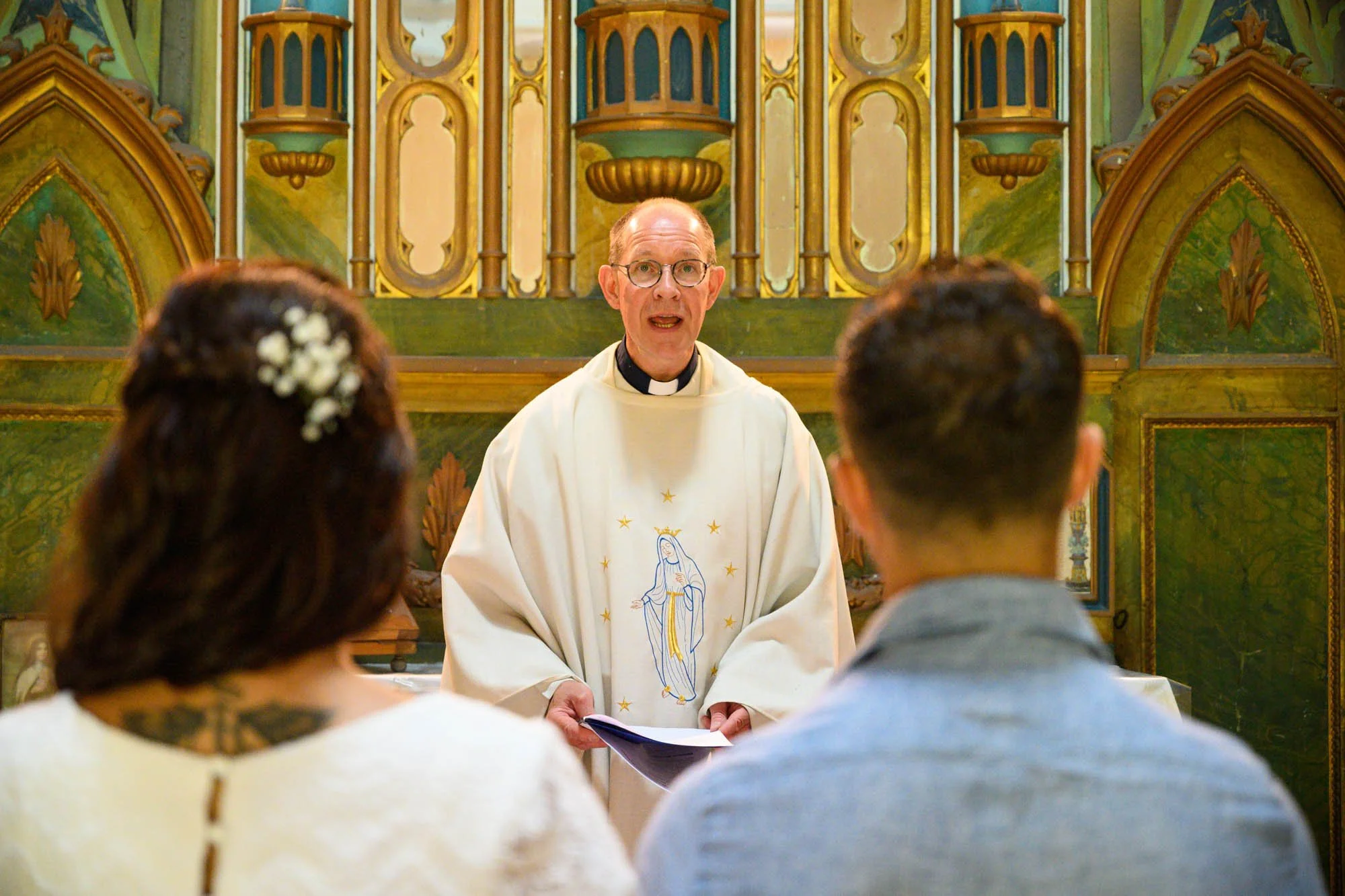 Un prêtre célèbre une cérémonie religieuse devant un couple assis dans une église ornée de décorations religieuses