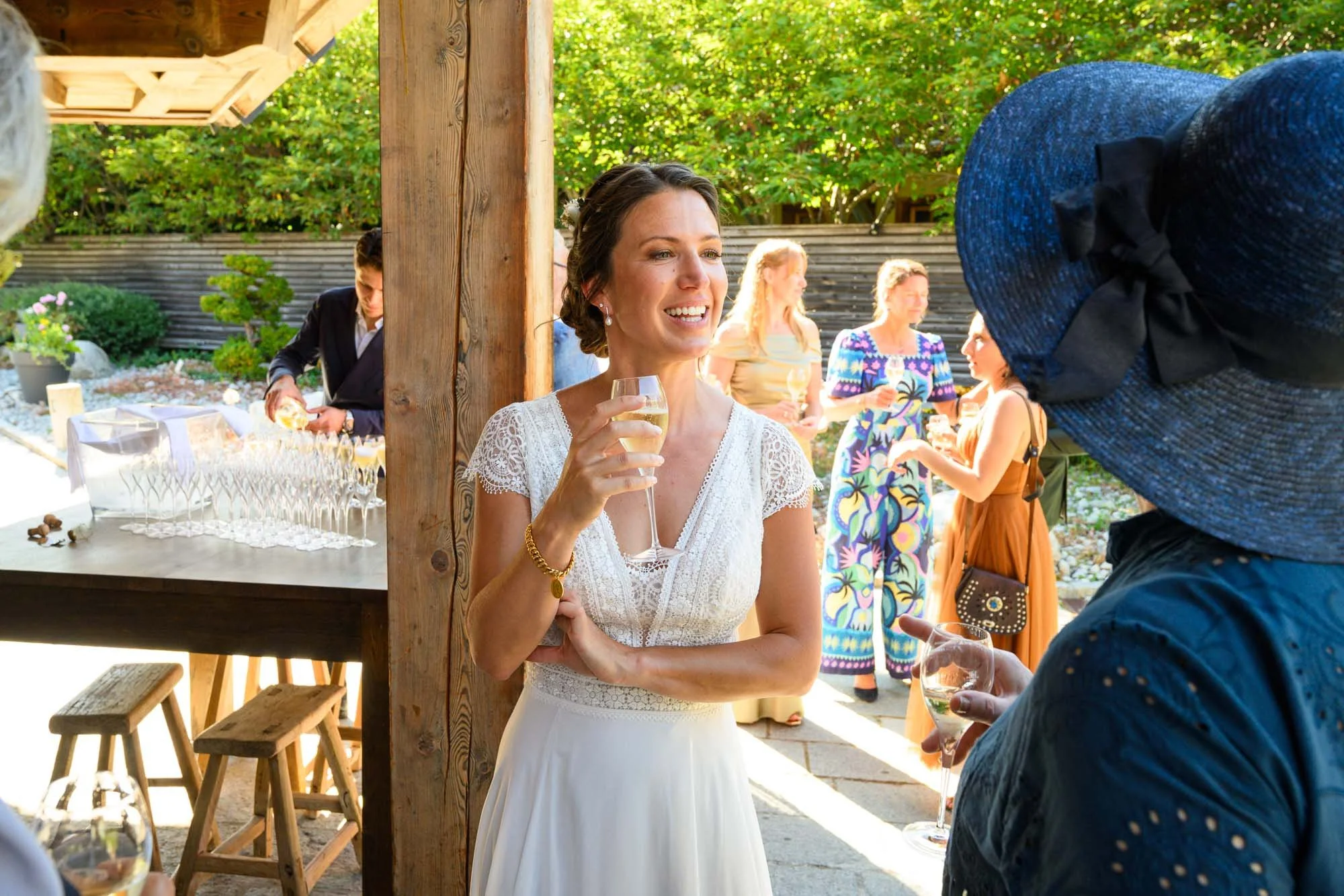 Une femme en robe blanche souriante, tenant un verre de champagne, parle à une autre personne portant un grand chapeau bleu lors d'une fête en extérieur.