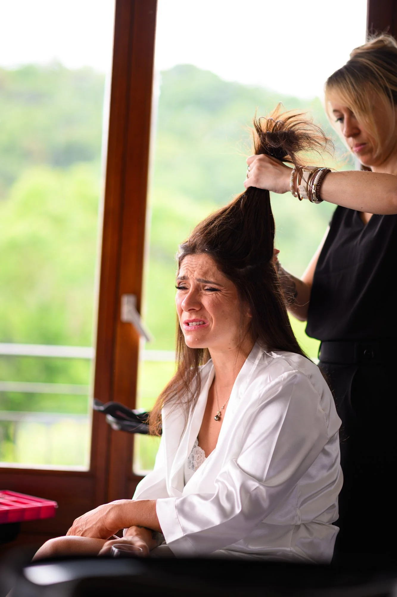 Une femme se faisant coiffer par une coiffeuse, avec une expression de gêne ou d'inconfort, à l'intérieur d'une pièce avec grande fenêtre et vue sur la nature.