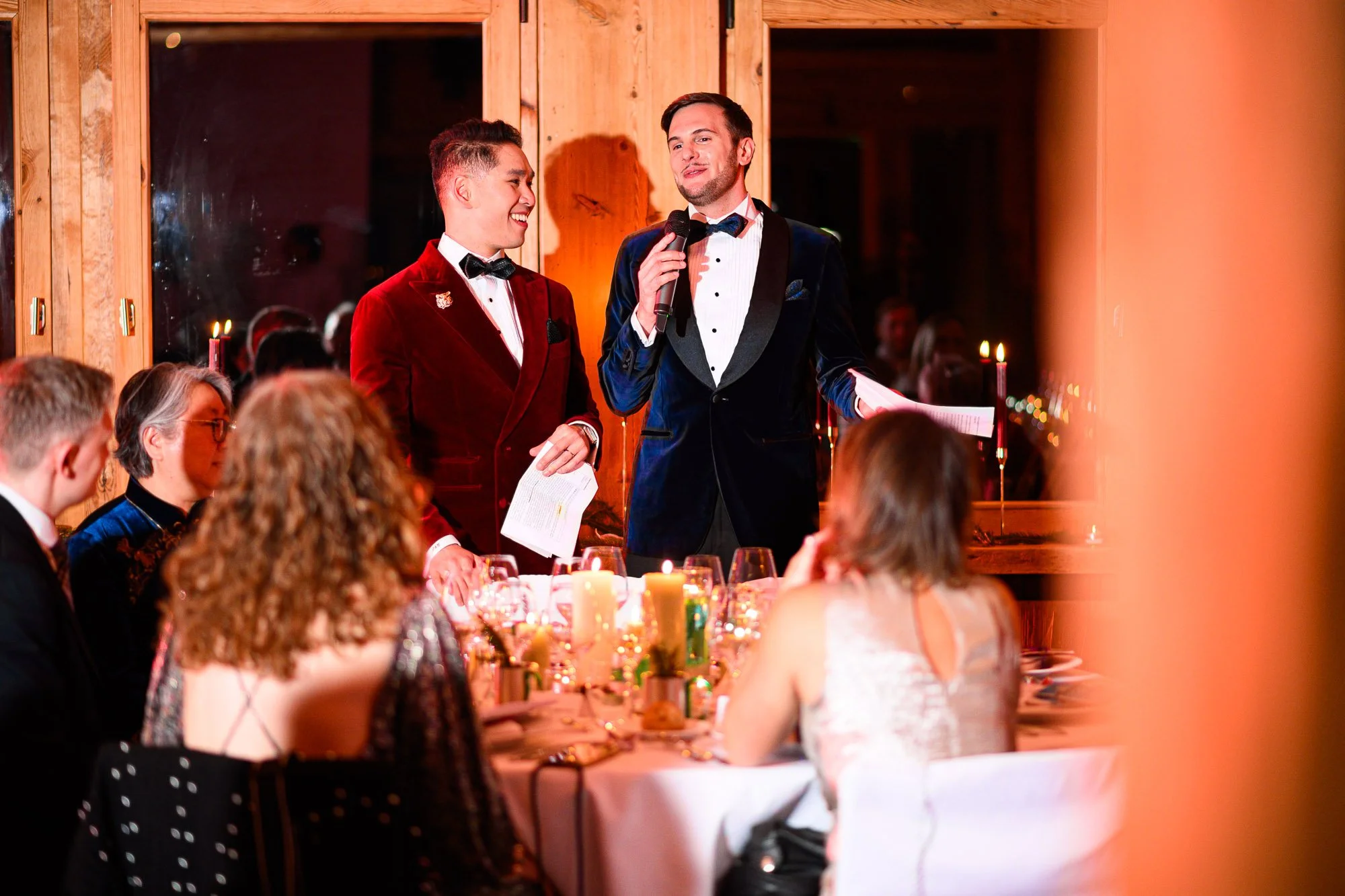 Deux hommes en costumes formels se tiennent devant une table lors d'un dîner ou d'une réception, avec un groupe de personnes assises autour d'eux, dans une ambiance chaleureuse et élégante.
