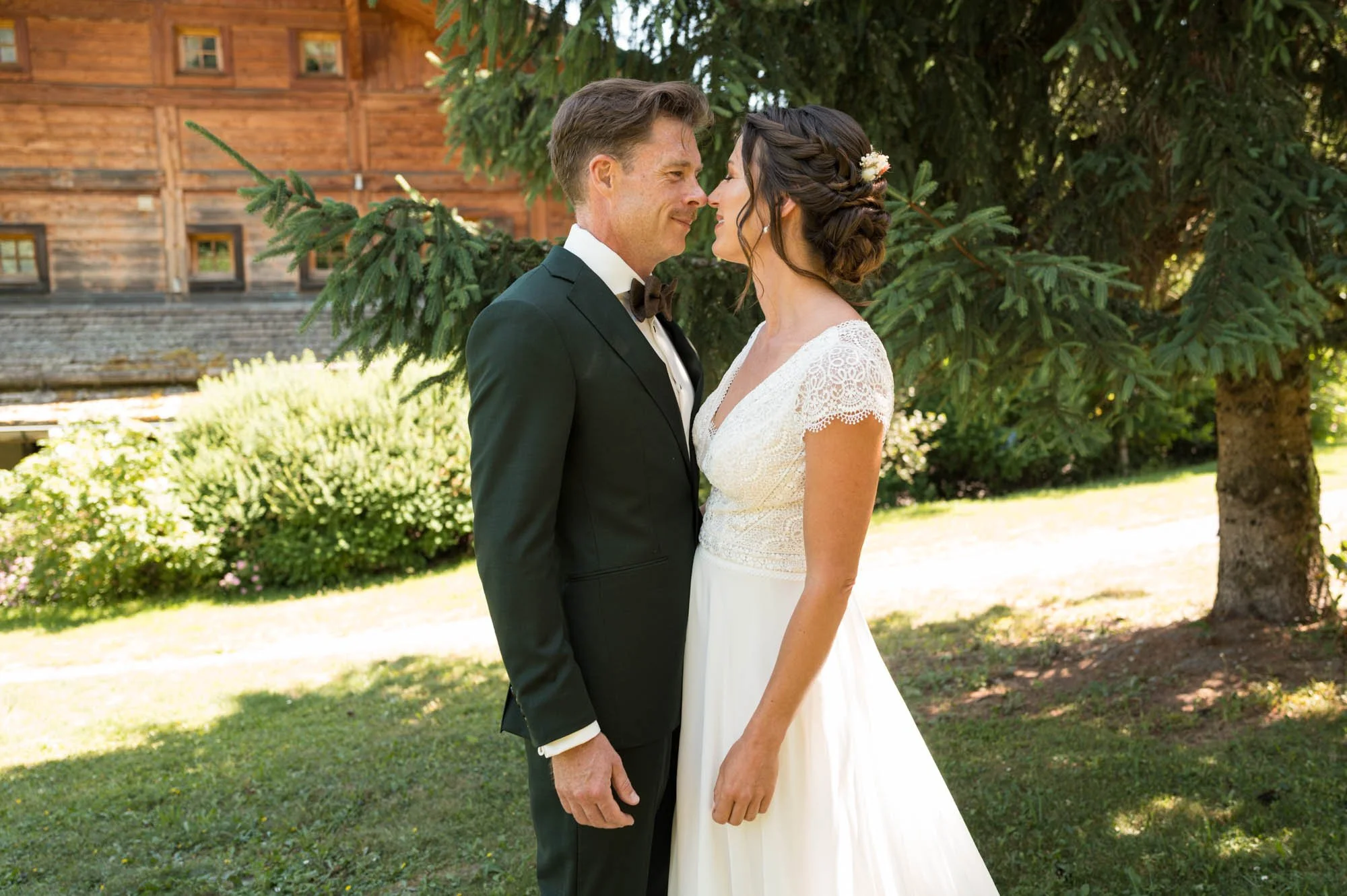 Un couple de mariés en vêtements de mariage s'embrassant devant un arbre en extérieur, avec une maison en bois en arrière-plan.