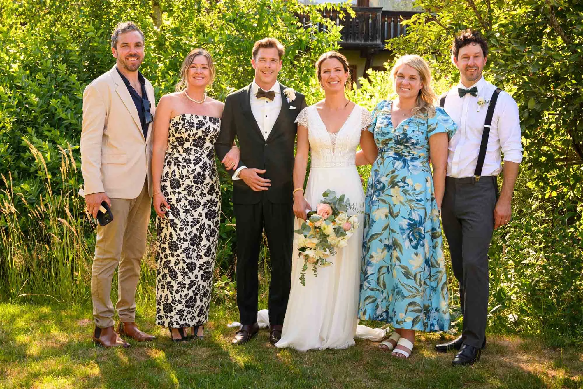 Groupe de sept personnes, dont un couple de mariés, posant en plein air dans un jardin ensoleillé, entouré de végétation verte. Le marié porte un costume noir avec une chemise blanche et un nœud papillon, la mariée porte une robe blanche avec un bouq