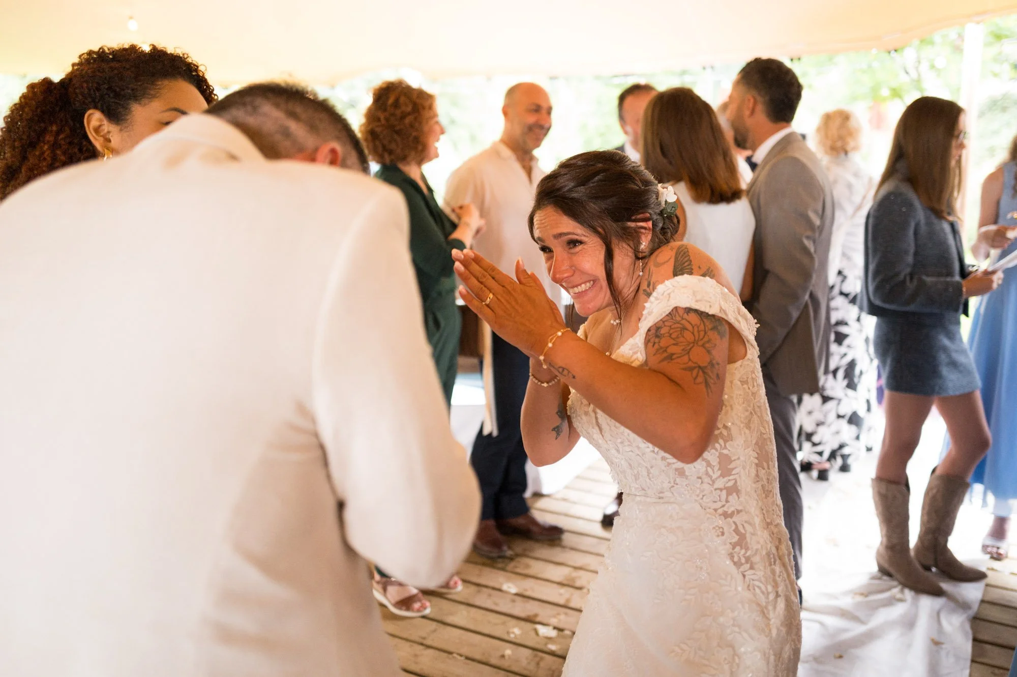 Une femme en robe de mariage souriante et émue échangeant un geste d'affection avec un homme lors d'une célébration, entourée de plusieurs personnes en habits élégants dans un cadre festif.