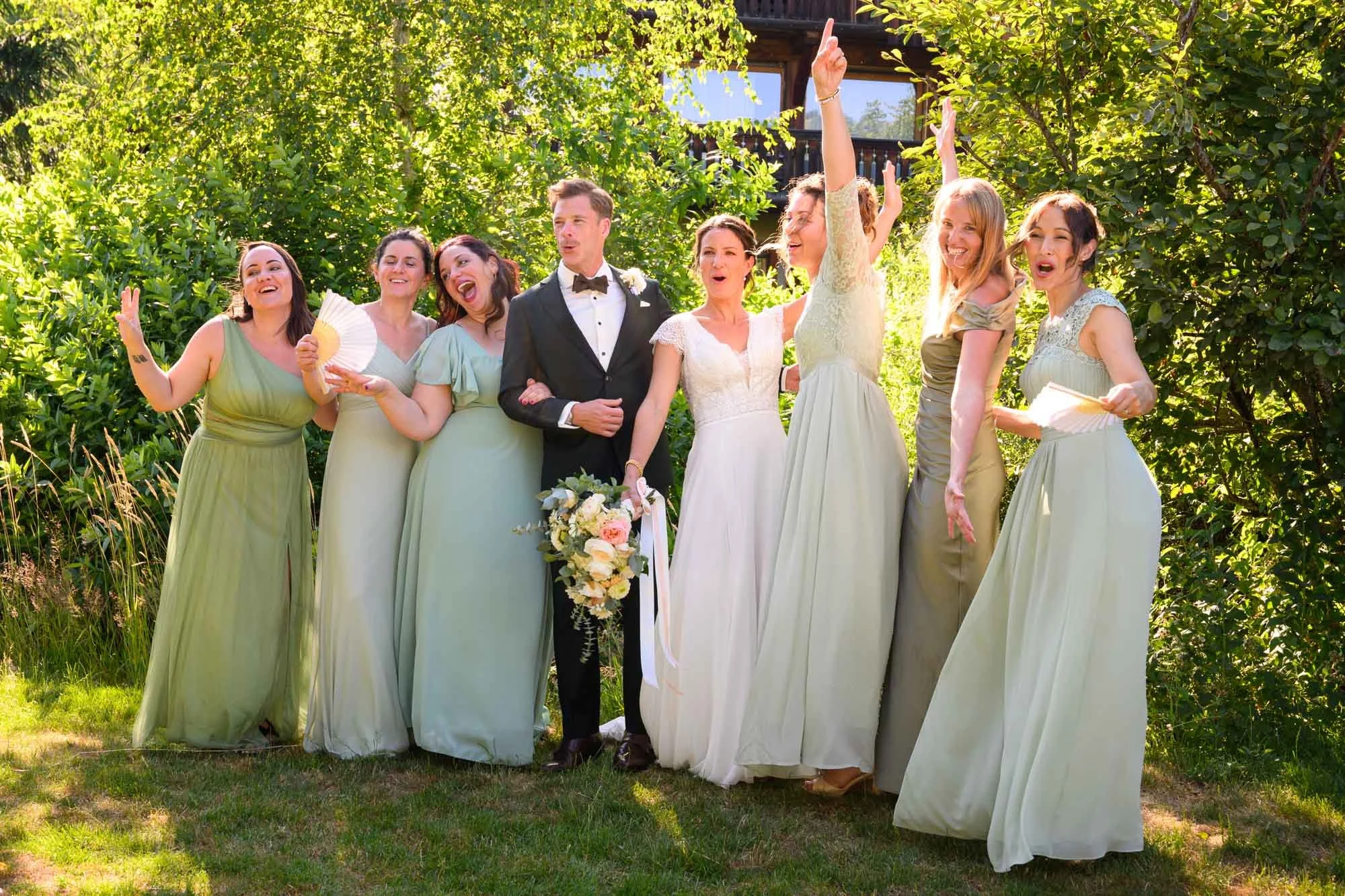 Groupe de personnes lors d'un mariage, comprenant le couple de mariés et des femmes en robes de cérémonie, se tenant dans un jardin avec des arbres en arrière-plan.