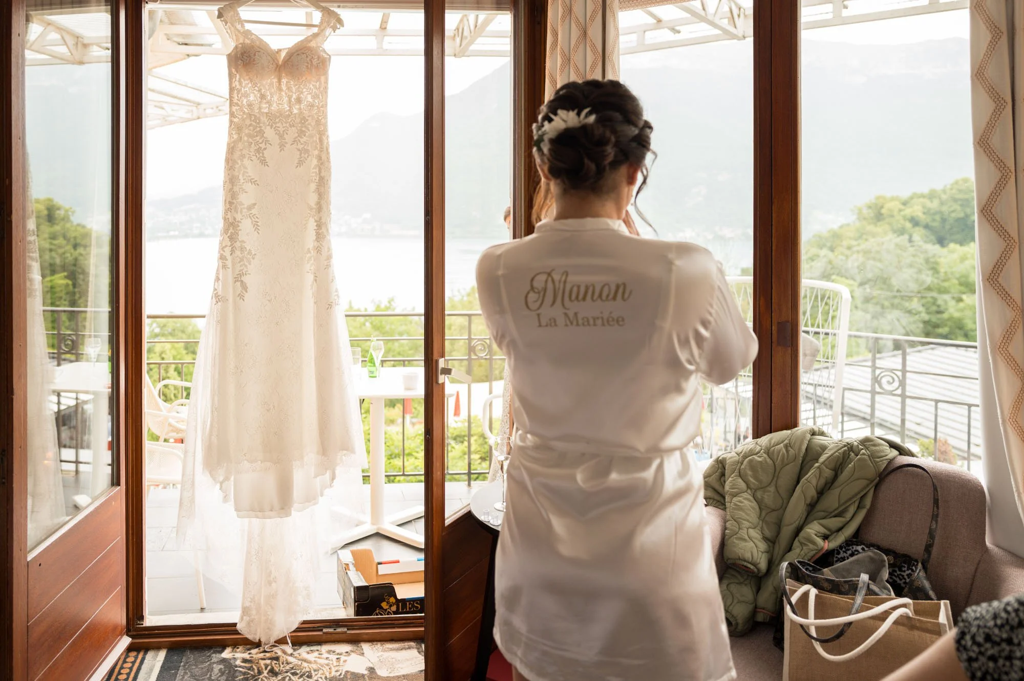 Une femme portant une robe de mariage de dos, derrière une robe de mariée suspendue à une fenêtre, avec un paysage de montagnes et un lac à l'extérieur.