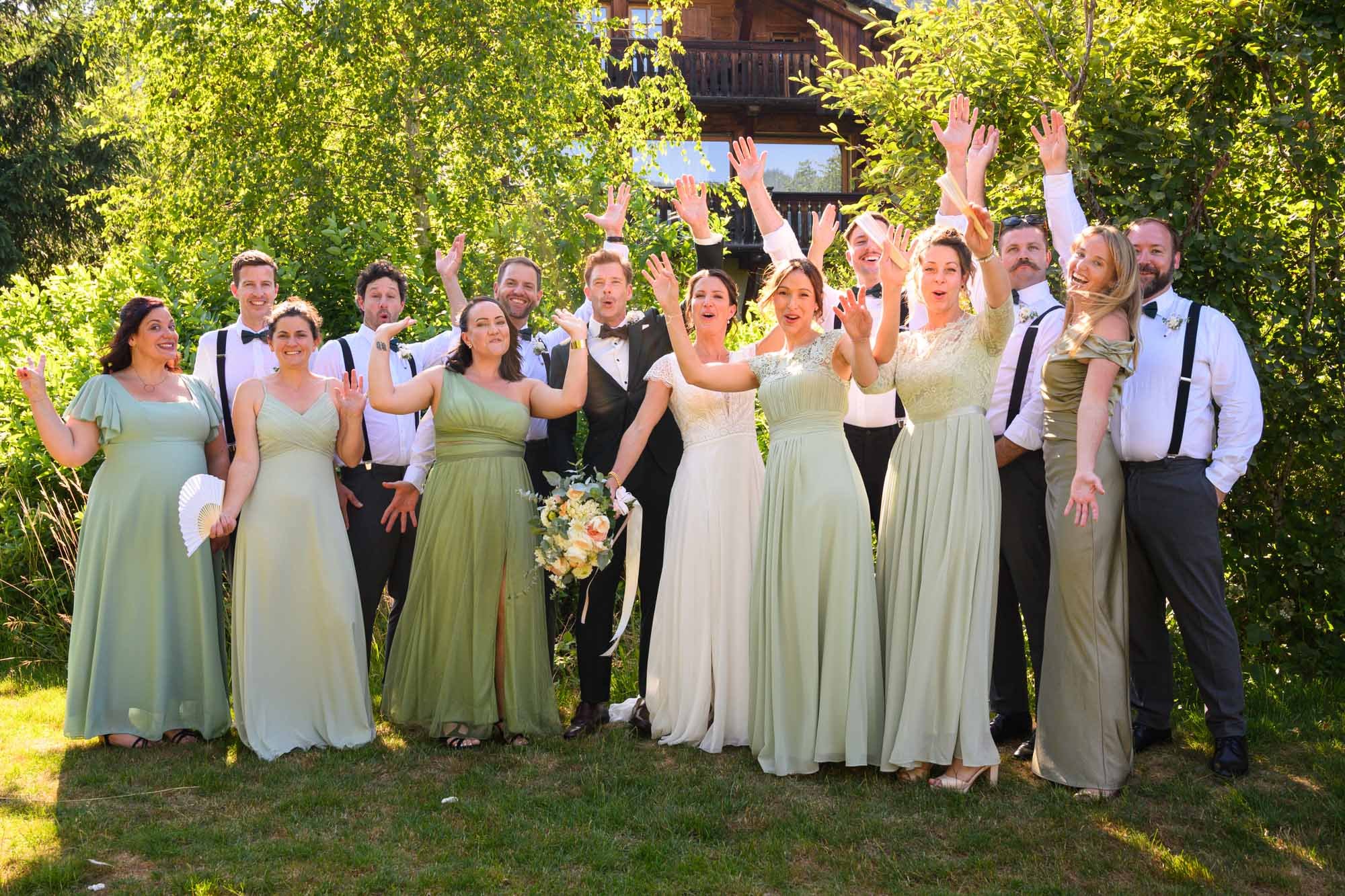 Groupe de personnes lors d'un mariage en plein air, avec femmes en robes vertes ou claires et hommes en chemises blanches et gilets noirs, en train de célébrer en levant les bras, entourés d'arbres verts et d'un bâtiment en bois en arrière-plan.