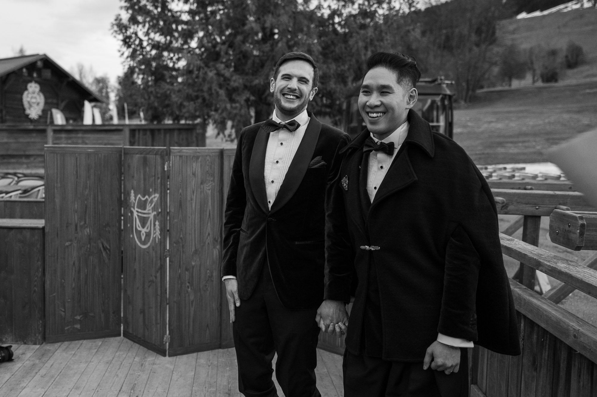 Deux hommes en smoking et manteau noir, tenant la main, sourient lors d'une occasion spéciale, en extérieur, avec un décor naturel en arrière-plan.