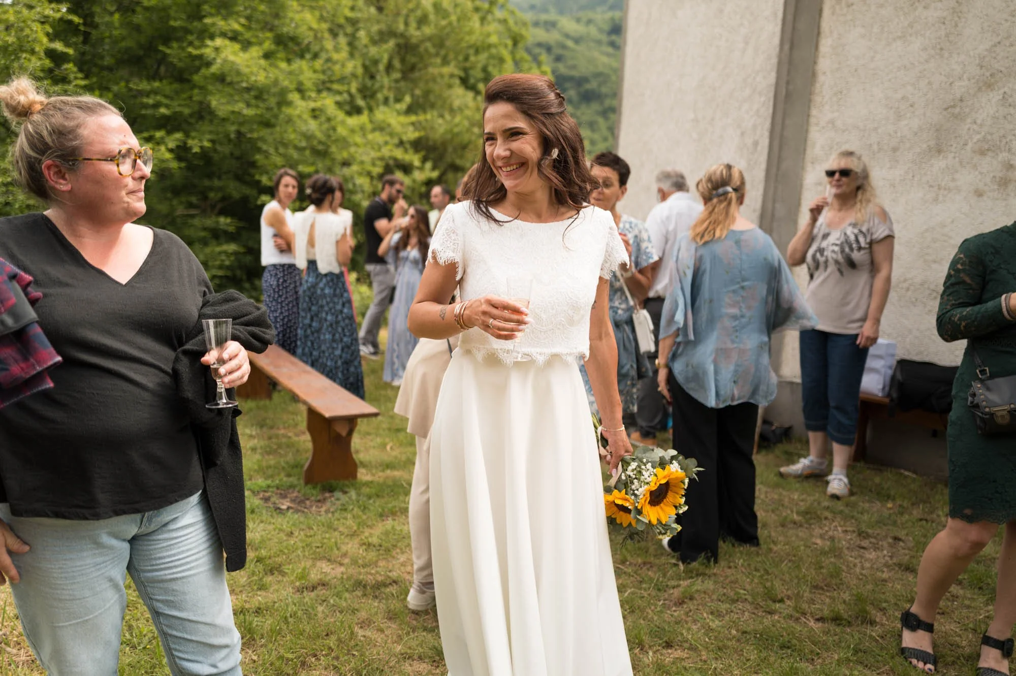 Une femme en robe de mariage tenant un bouquet de tournesols et un verre, souriant lors d'une fête en plein air avec plusieurs personnes en arrière-plan.