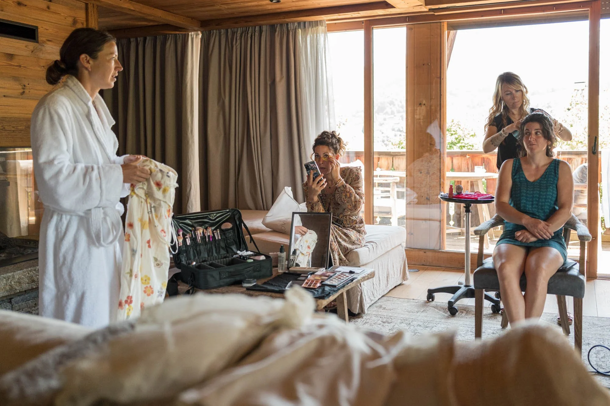 Jeune femme en robe verte assise dans une pièce en bois, en train d'être coiffée par une coiffeuse. À gauche, une autre femme en peignoir blanc montre du maquillage. Sur la table devant elles, une trousse de maquillage et un miroir.