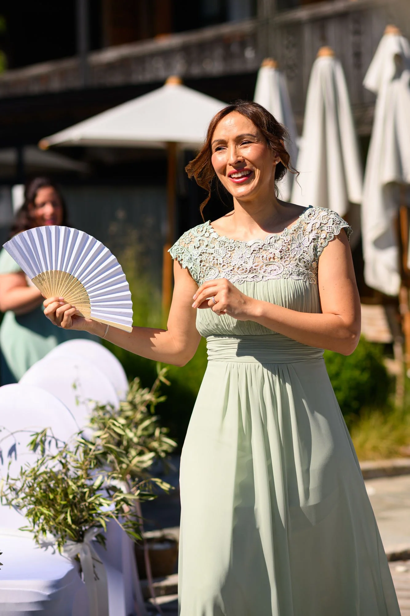 Une femme souriante en robe beige claire et dentelle, tenant un éventail, lors d'une cérémonie en plein air sous un soleil brillant.