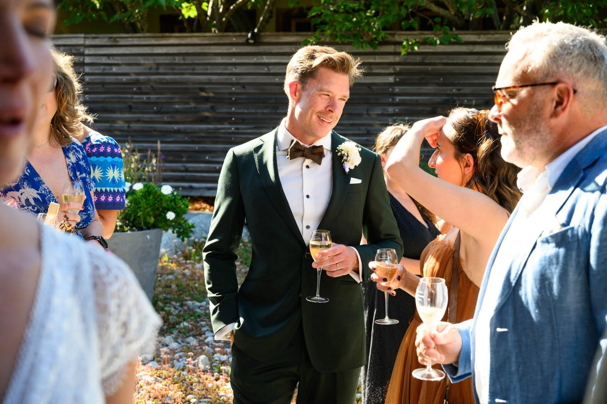 Groupe de personnes en costume et tenues élégantes, célébrant lors d'un mariage en extérieur avec verres de champagne, soleil et décoration florale
