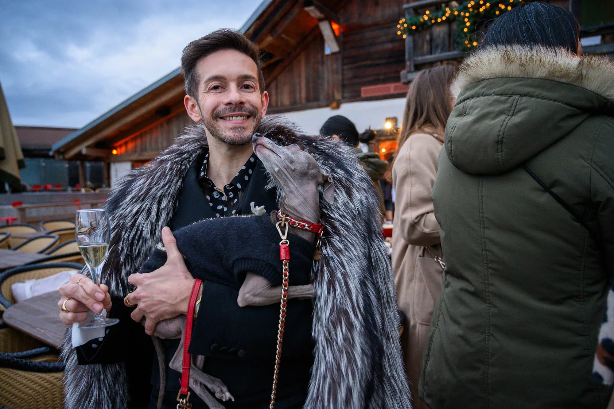 Un homme souriant tenant un verre de champagne et un chien avec le visage proche de lui lors d'une fête en extérieur avec des personnes autour