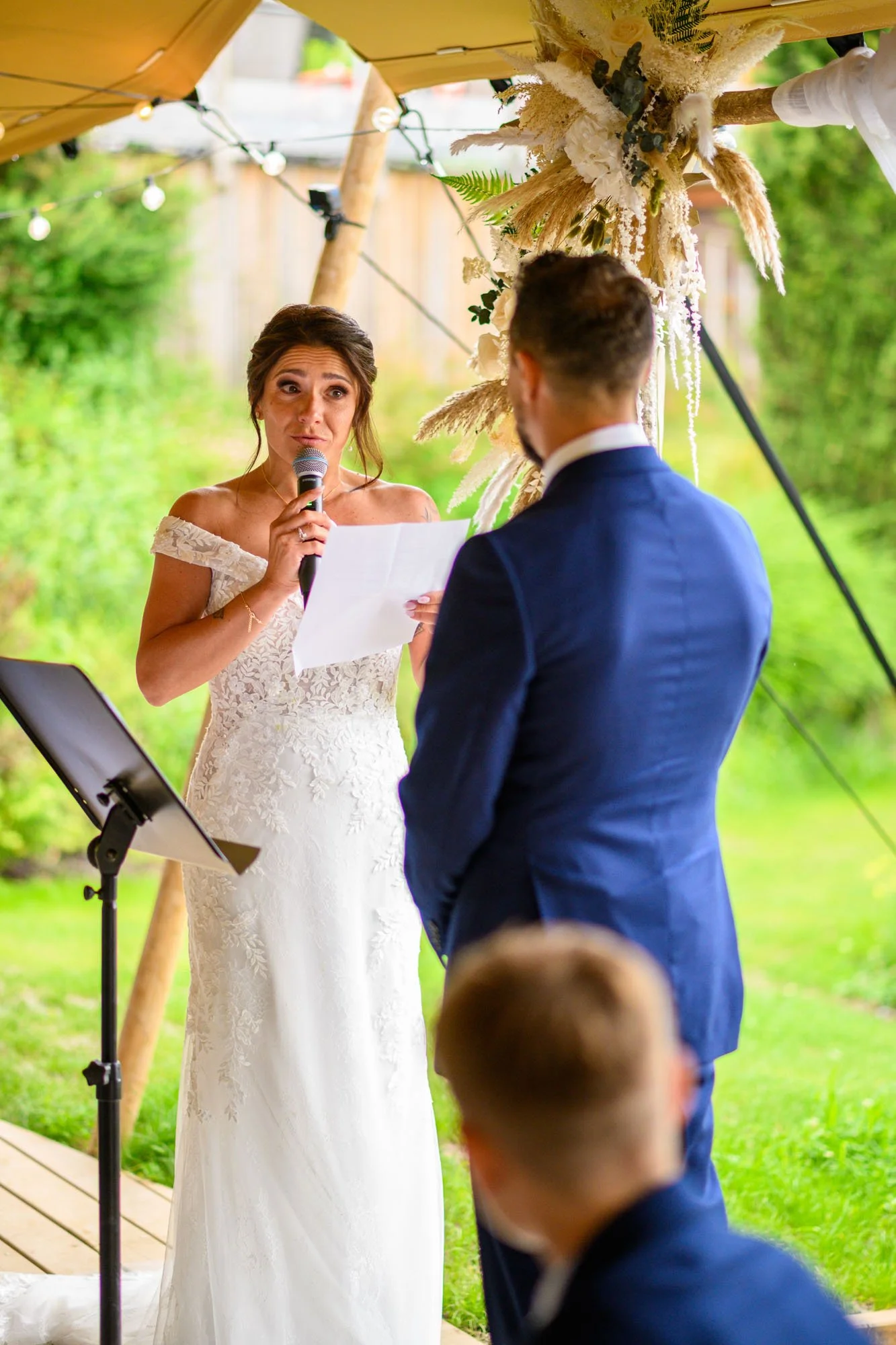Une cérémonie de mariage en plein air avec une femme en robe de mariée lisant pendant la cérémonie, un homme en costume bleu écoutant, et un enfant en avant-plan. Décoration florale et guirlandes lumineuses sont visibles.