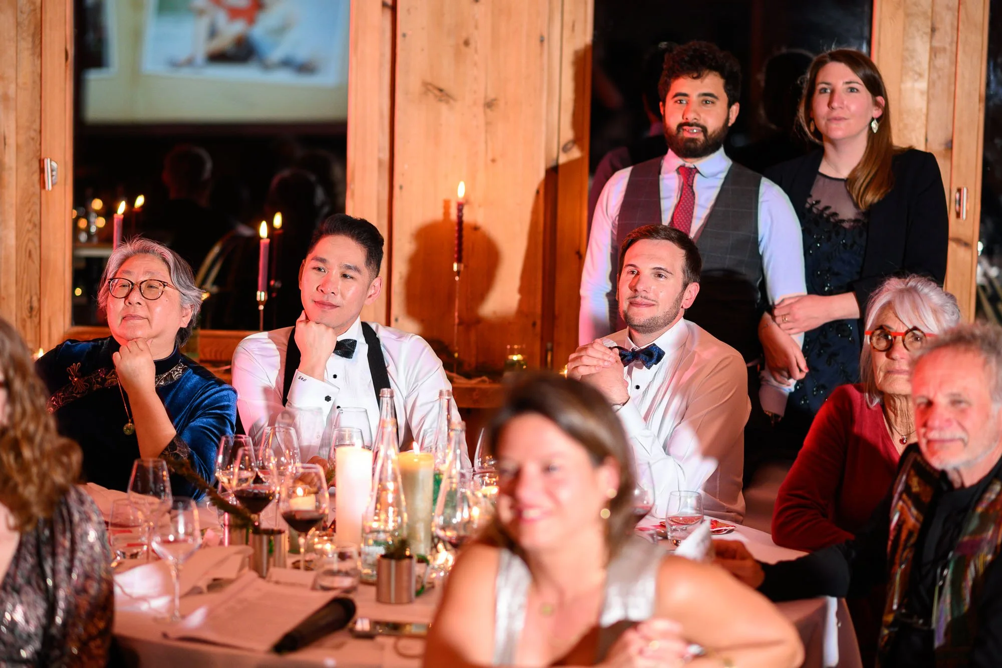 Groupe de personnes assises et debout lors d'une célébration ou d'un dîner, dans un cadre chaleureux avec bougies et décor en bois.