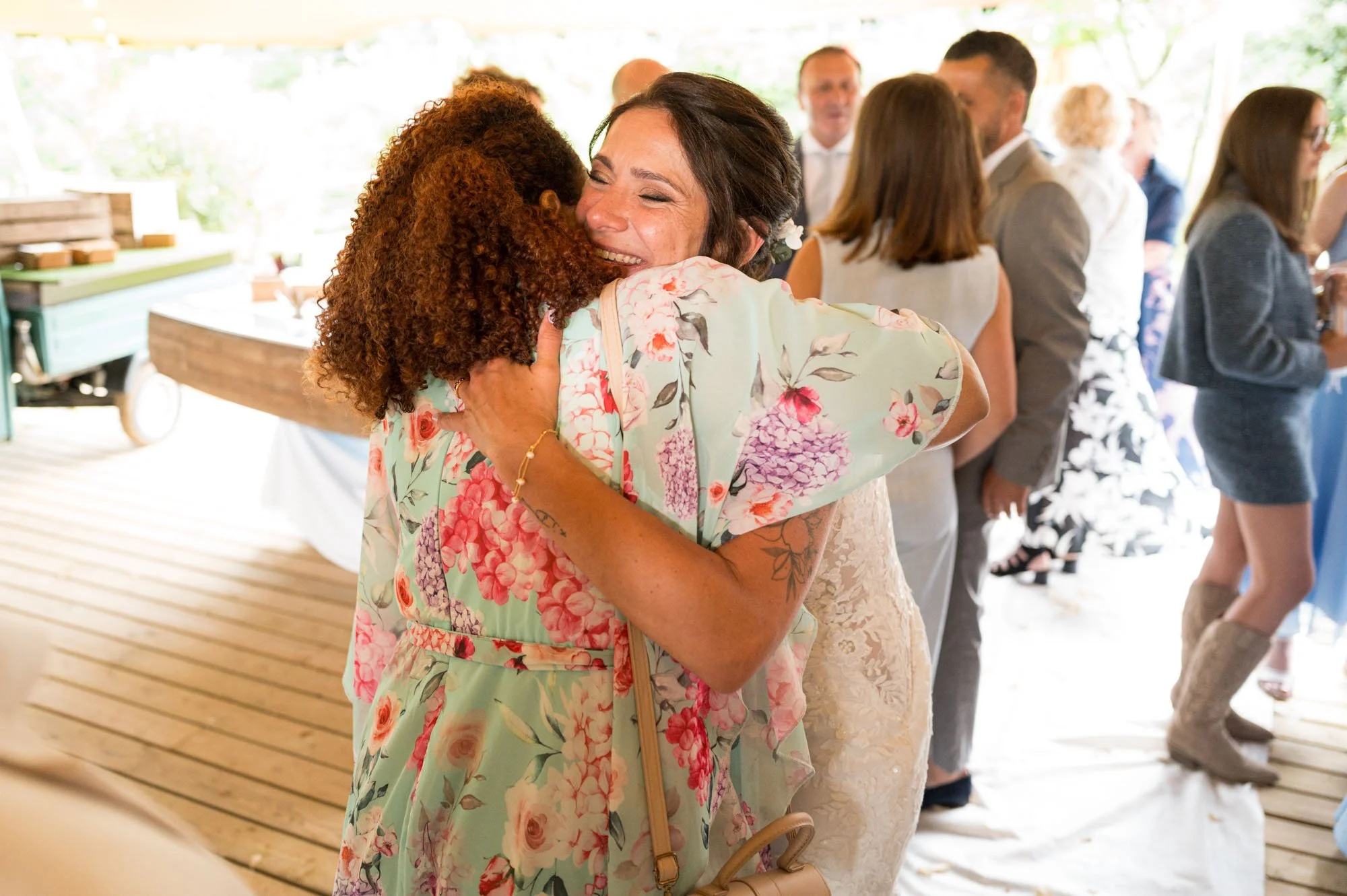 Deux femmes s'embrassent chaleureusement lors d'une célébration, entourées d'autres personnes qui socialisent dans un espace lumineux et accueillant.