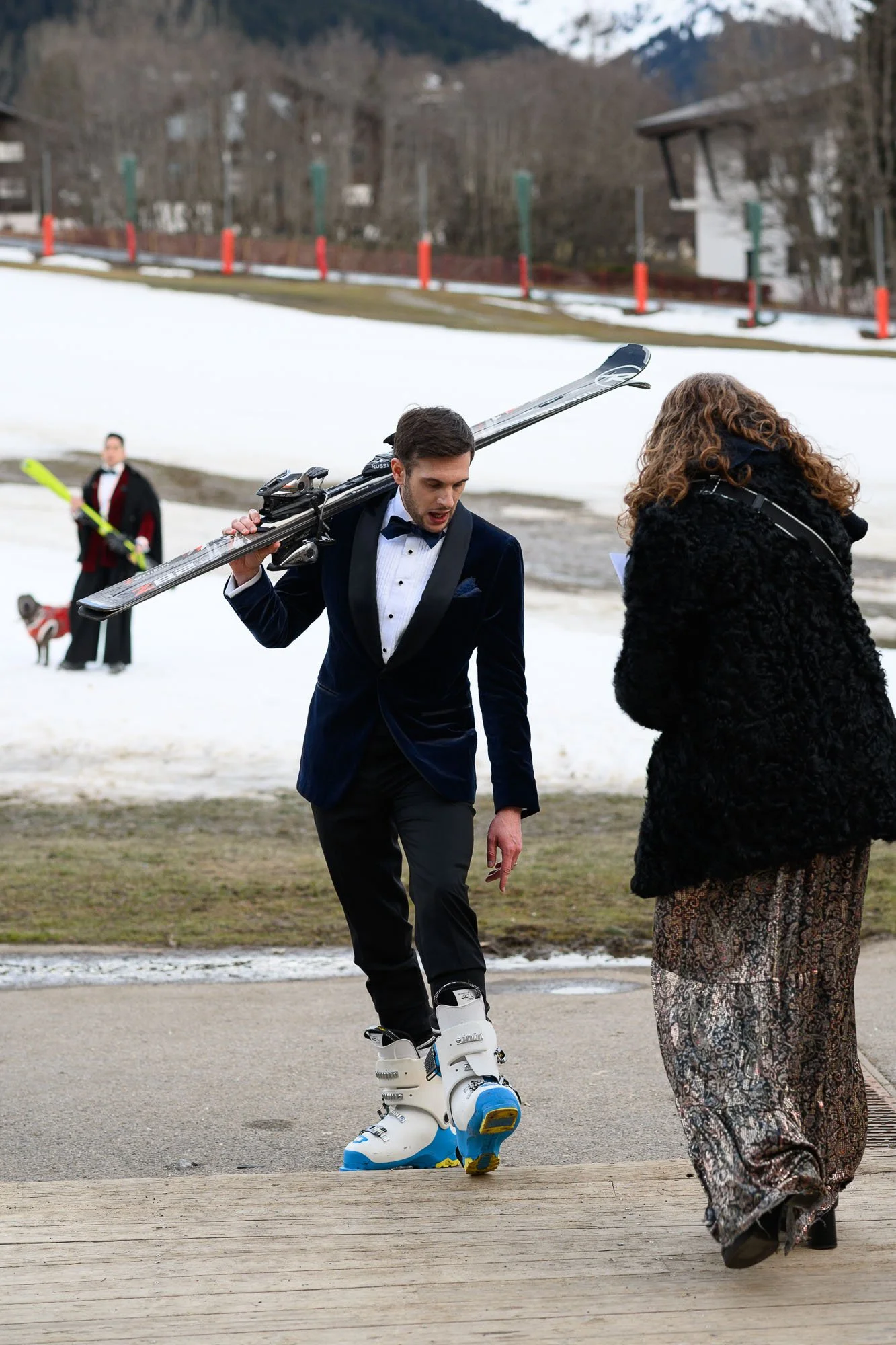 Un homme en smoking avec des skis et des bottes de ski, parlant avec une femme portant un manteau noir et une longue jupe, dans un paysage enneigé.