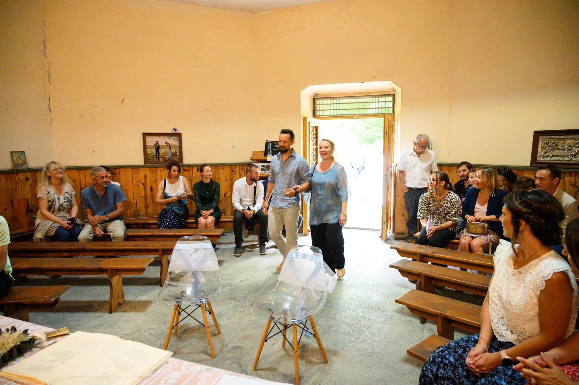 Groupe de personnes assises dans une salle, certaines souriantes, d'autres regardant vers la scène, avec deux personnes marchant vers le centre, à l'entrée d'une pièce éclairée par la lumière naturelle.