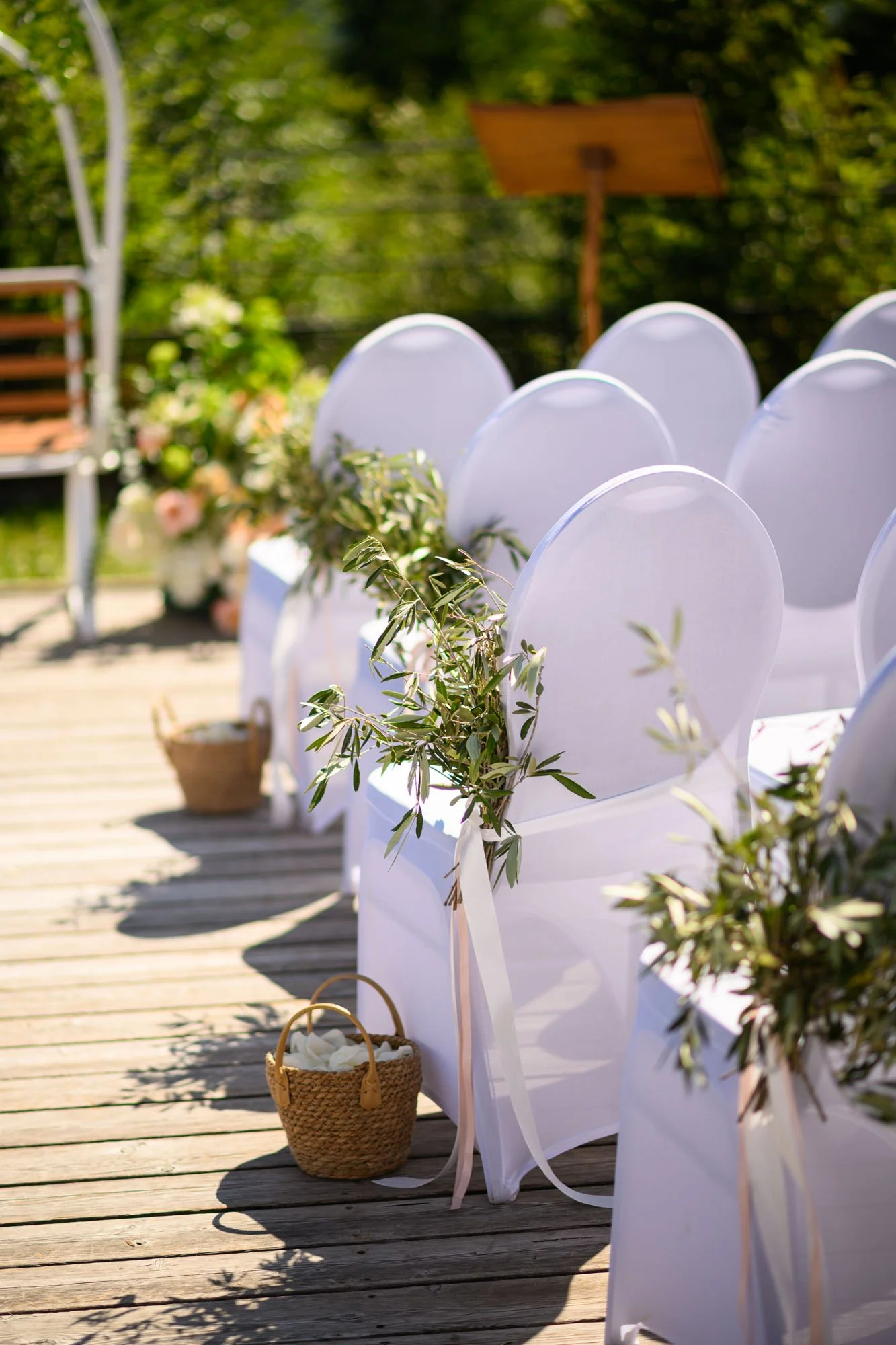 Chaises blanches décorées avec des rubans et des branches d'olivier dans un cadre extérieur pour une cérémonie, probablement un mariage, sur une terrasse en bois en plein air avec un fond de verdure.