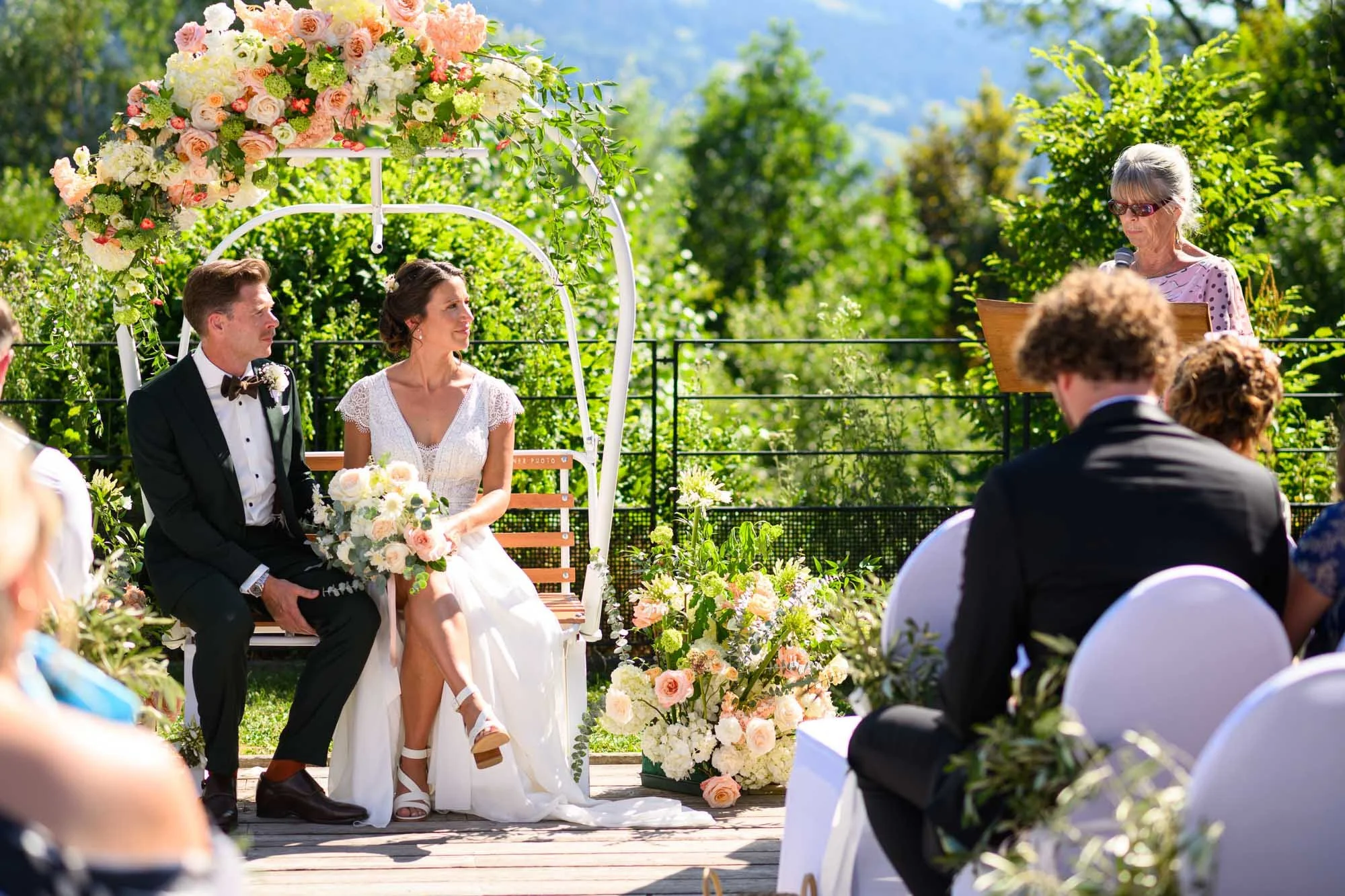 Un couple de mariés lors de leur cérémonie de mariage en plein air, assis sur un banc sous une arche décorée de fleurs, avec un officiant lisant ses vœux, entourés de convives dans un jardin ensoleillé.