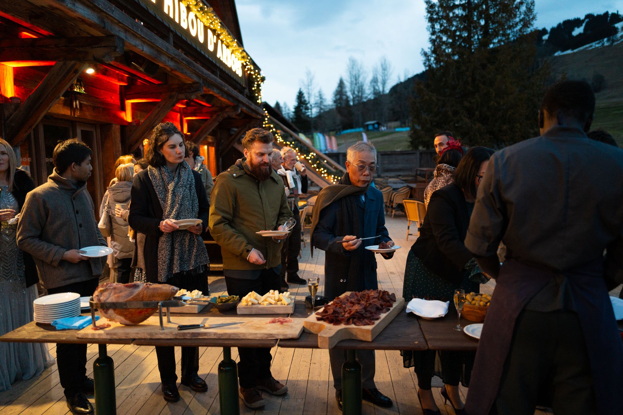 Groupe de personnes faisant la queue pour un buffet de charcuterie, fromage et autres plats lors d'un événement en extérieur en soirée, avec un bâtiment en bois décoré de guirlandes lumineuses en arrière-plan.