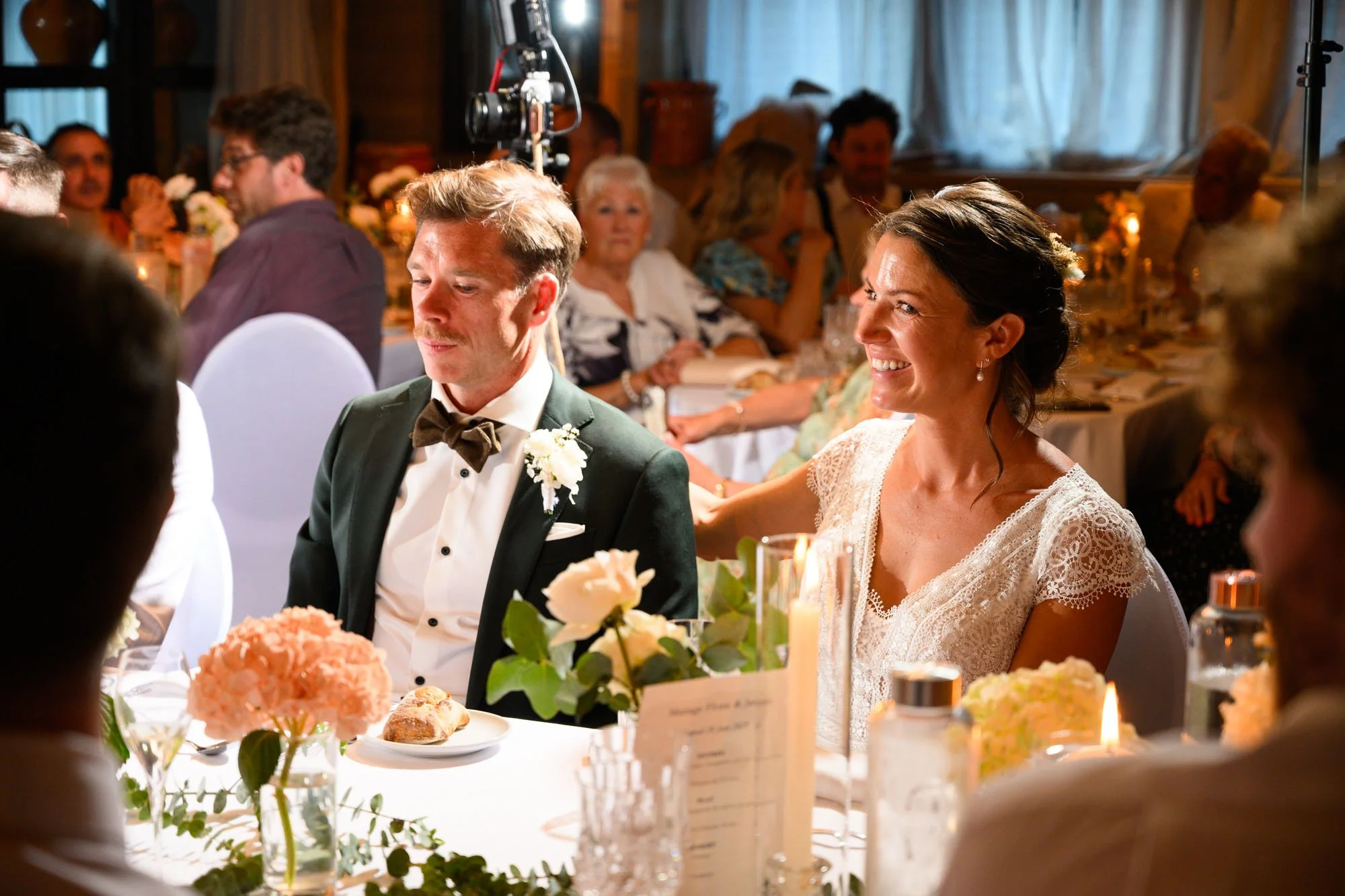 Mariée et marié assis à une table lors d'un mariage, entourés d'invités, dans une salle décorée avec des fleurs et des bougies.