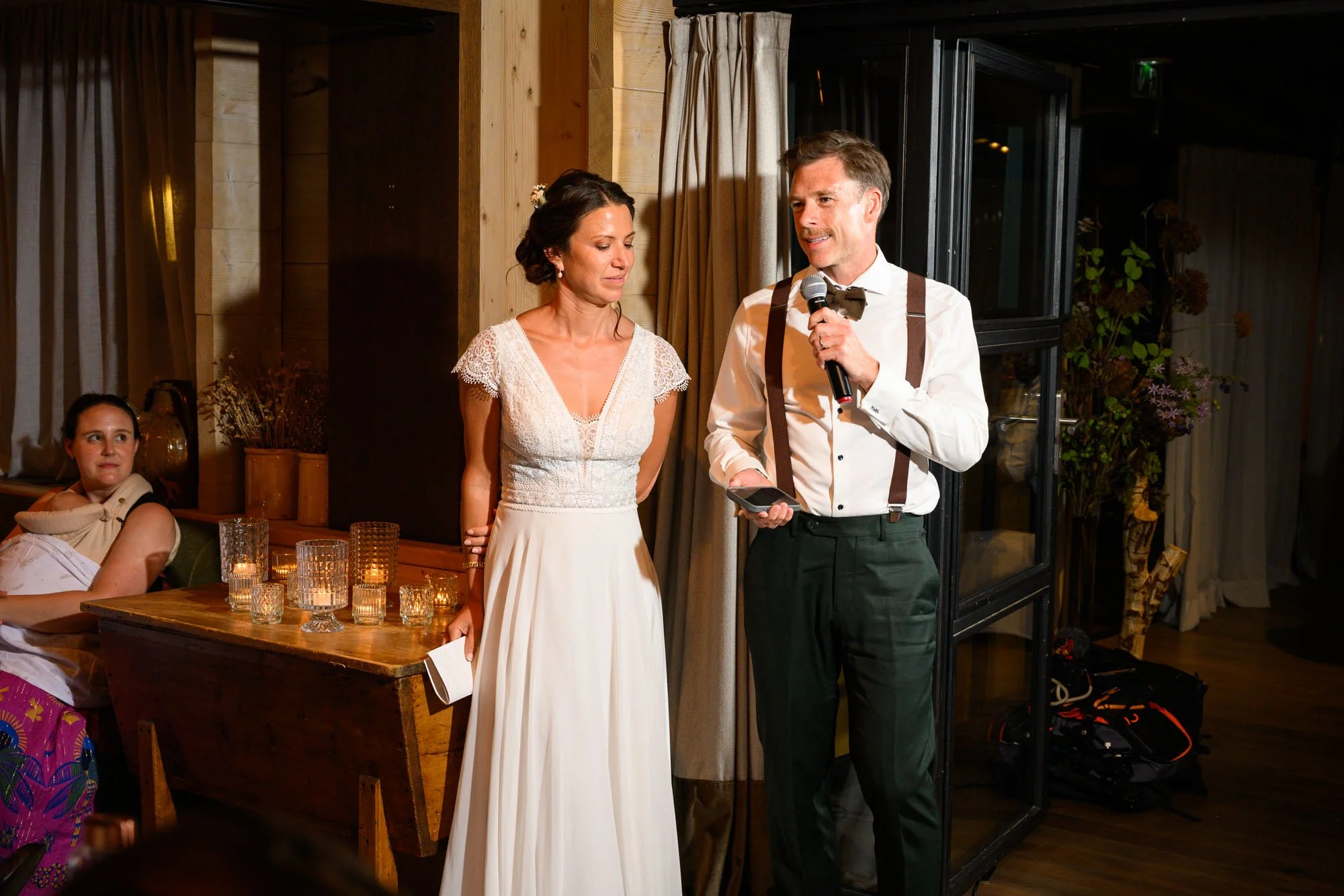 Un homme et une femme en costume de mariage donnant un discours lors d'une cérémonie, avec une femme assise à gauche devant une table avec des bougies.