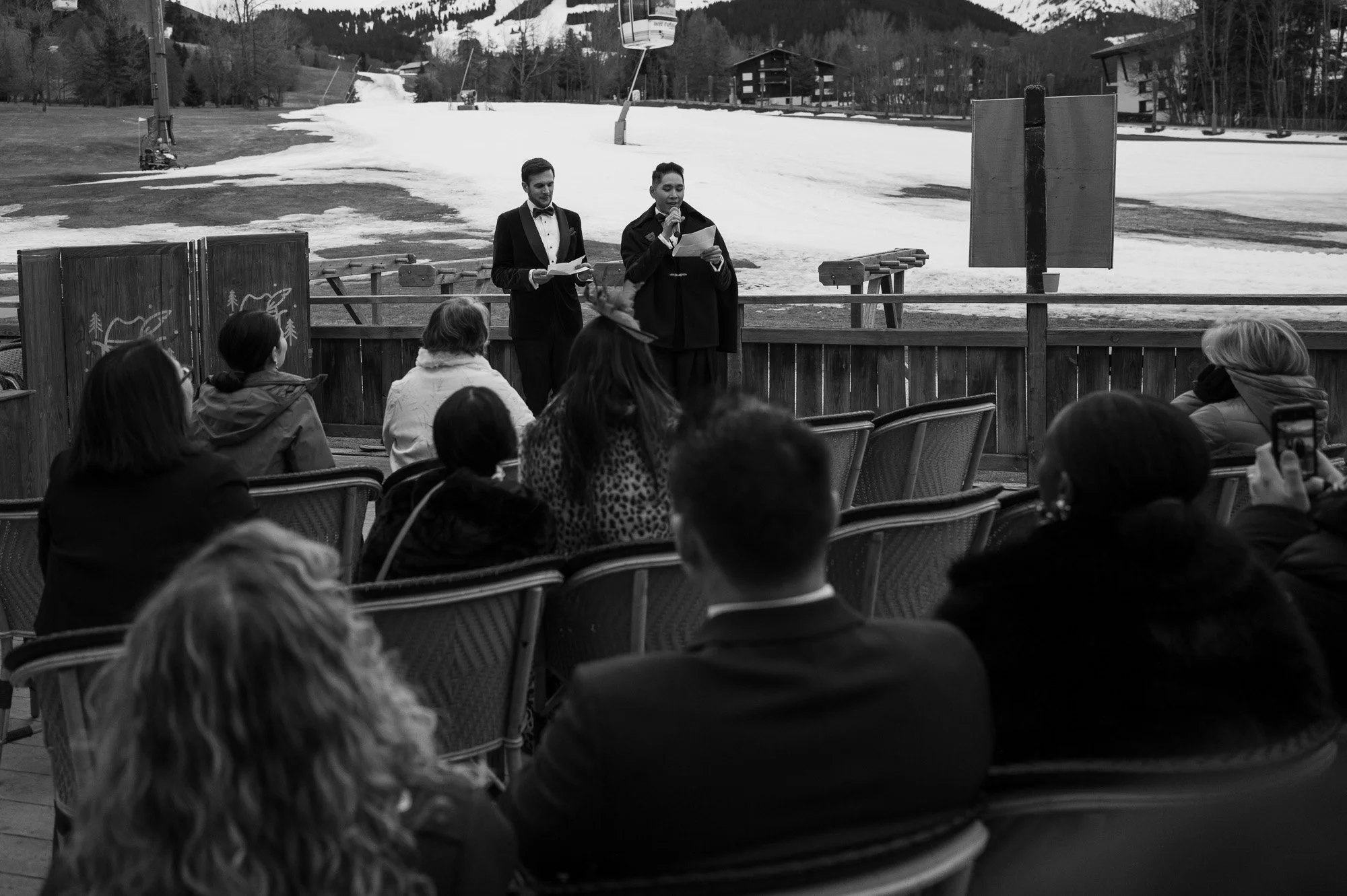 Un groupe de personnes assises regardant un spectacle en plein air avec deux hommes sur scène, en costumes, parlant à un micro, avec un décor enneigé en montagne en arrière-plan.
