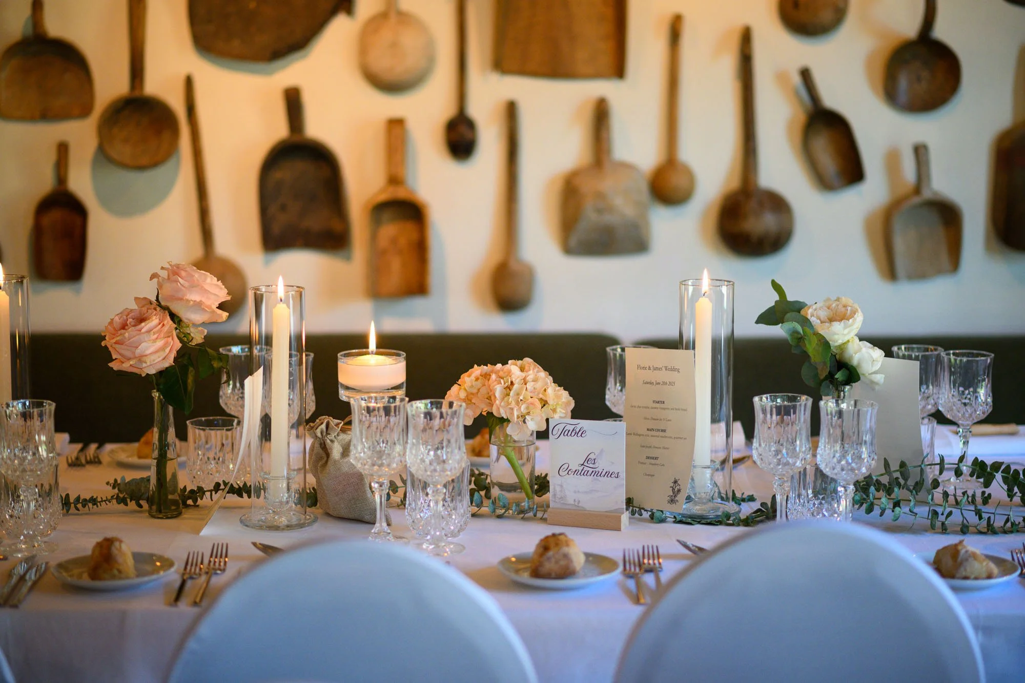 Table de mariage décorée avec des fleurs roses et blanches, des bougies, des verres en cristal, et un menu, dans un cadre rustique avec des outils en bois suspendus au mur.